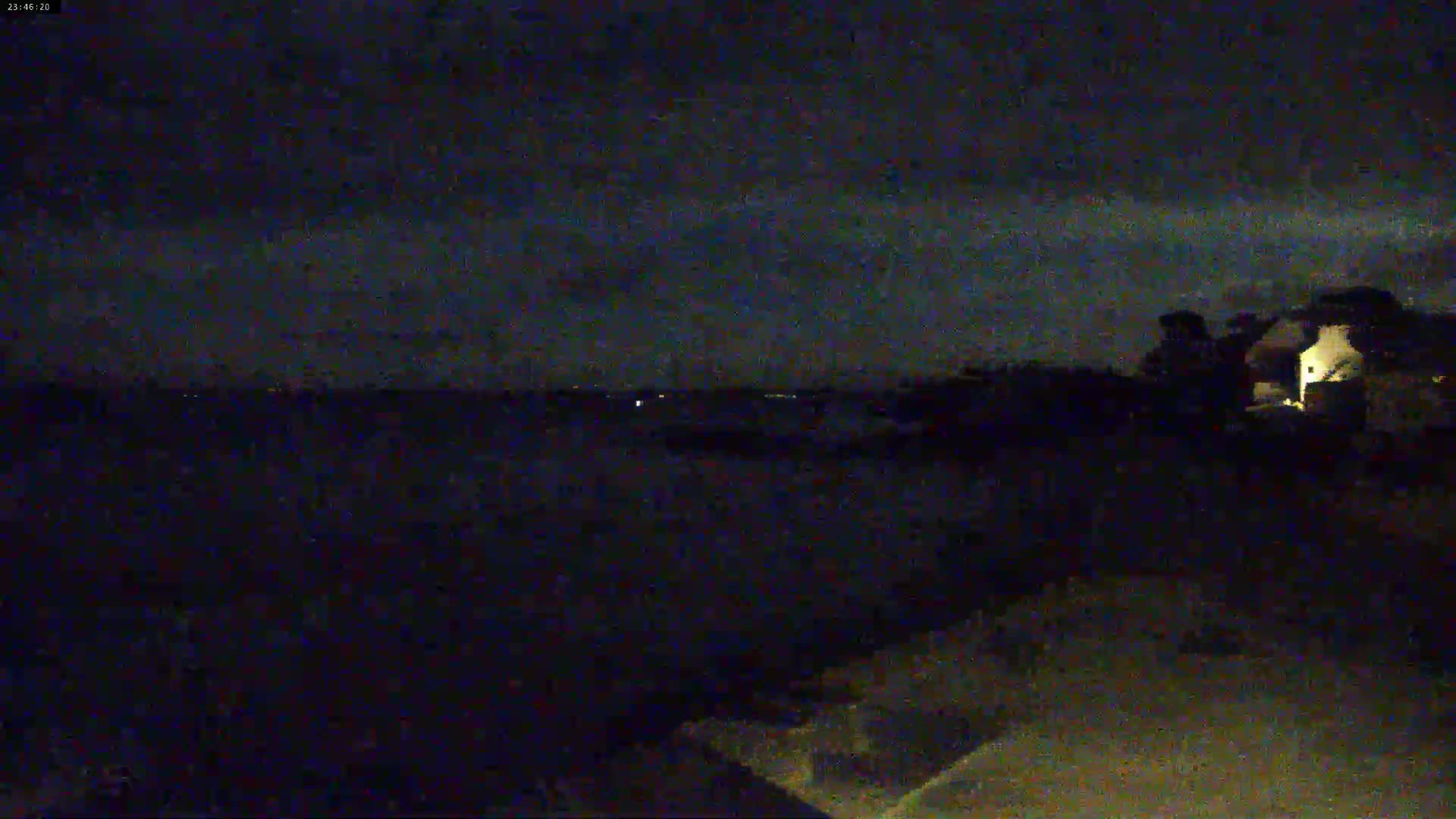 A nighttime view of a dark landscape with a faintly lit building in the distance under a mostly clear sky.