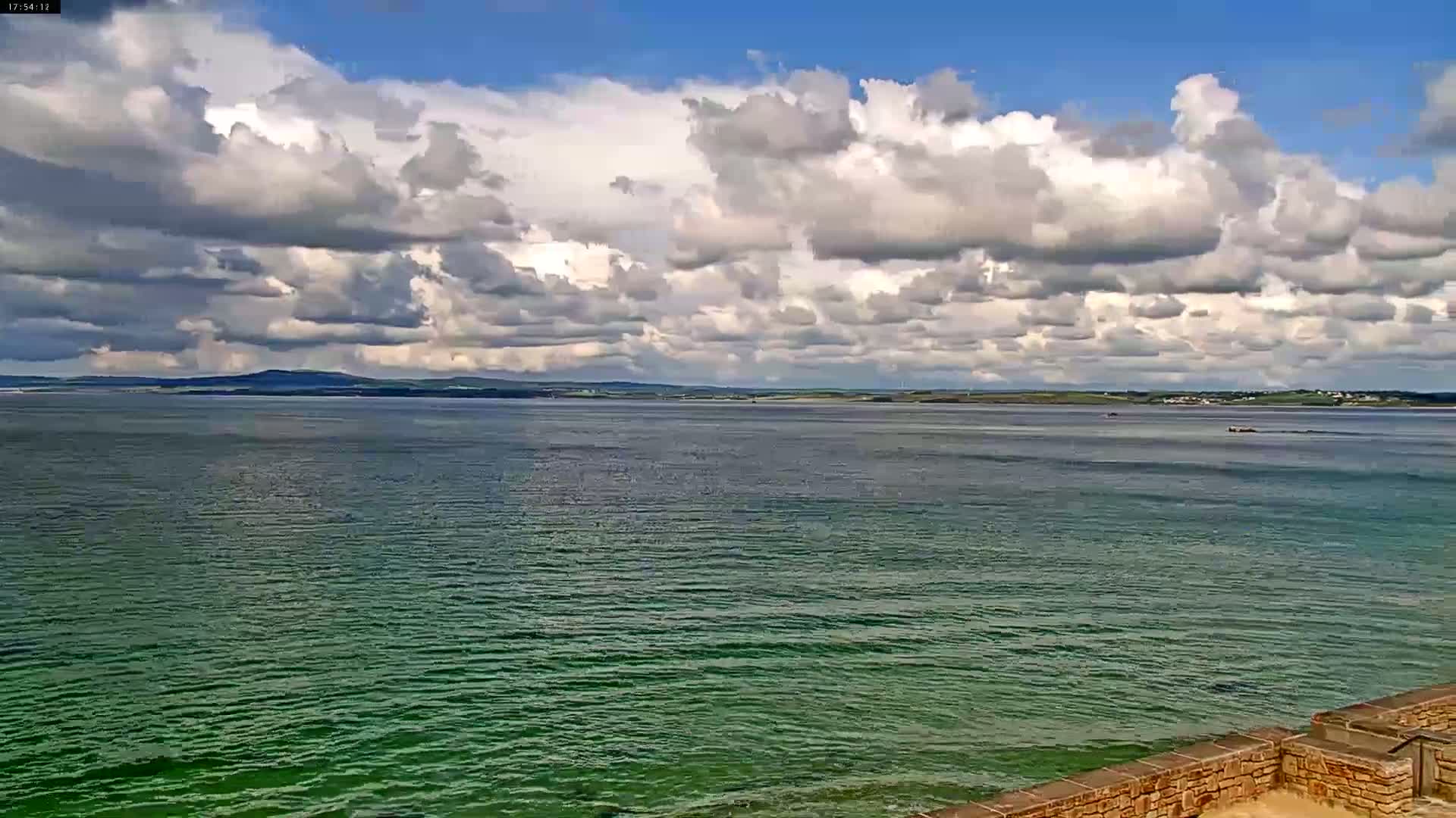 A partly cloudy sky overlooks a calm, green-tinged body of water, with distant land visible on the horizon.