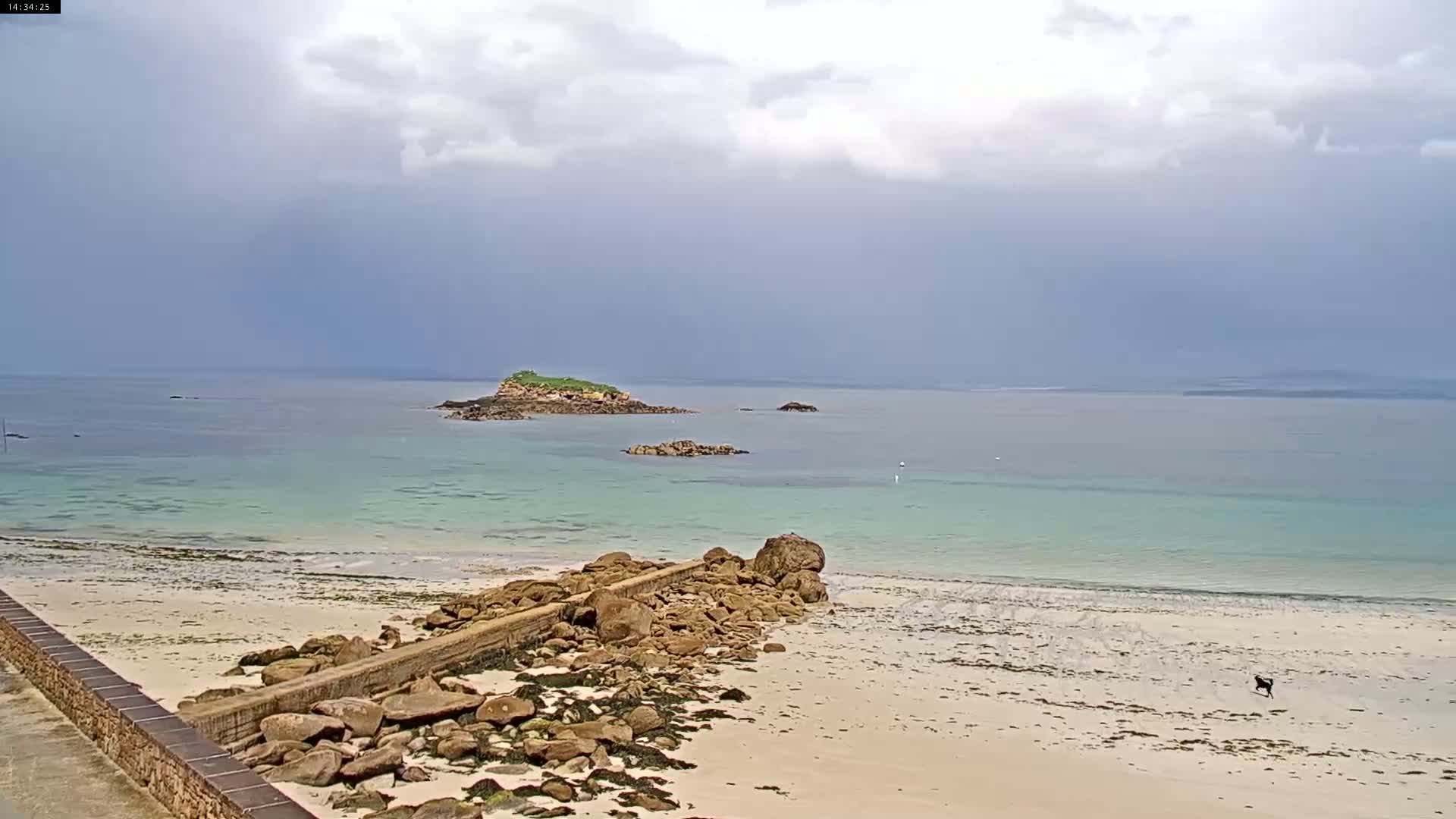 A sandy beach with a rocky jetty stretches toward a calm, light-green sea under a cloudy sky, with several small islands visible in the distance.