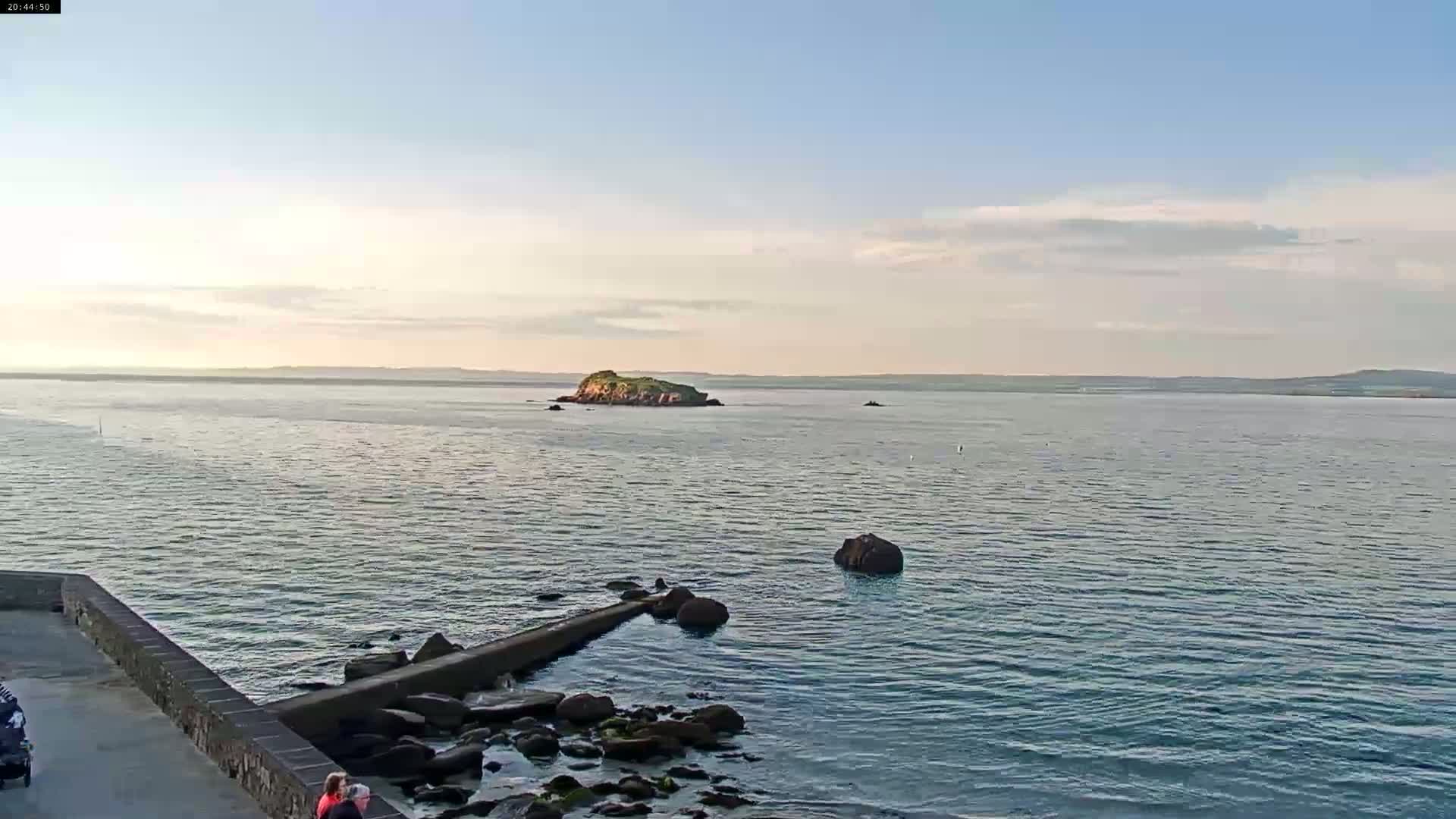 A calm body of water under a partly cloudy sky features a small, green island in the distance, a rocky shoreline, and two people near a low wall.
