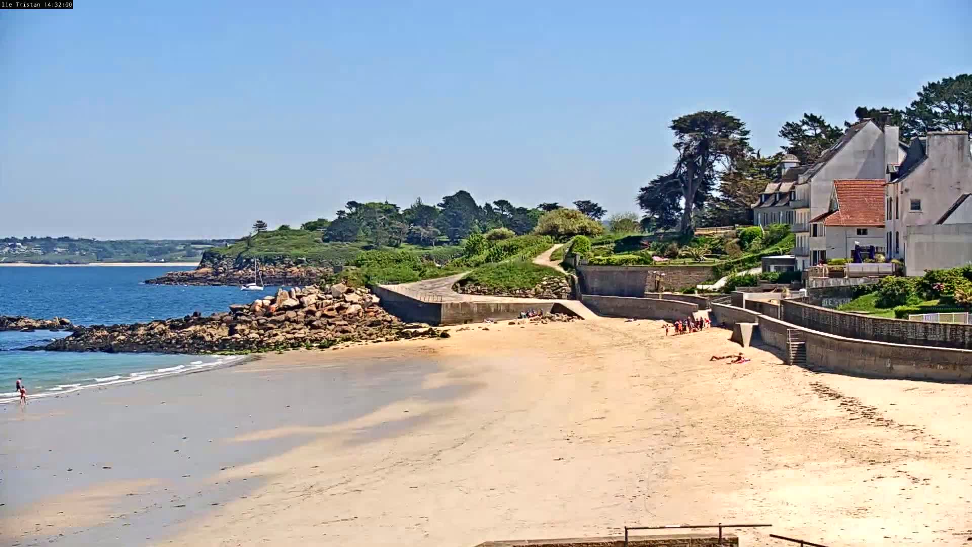 A sandy beach with people sunbathing and a few people in the water, backed by a rocky coast and several houses on a sunny day.