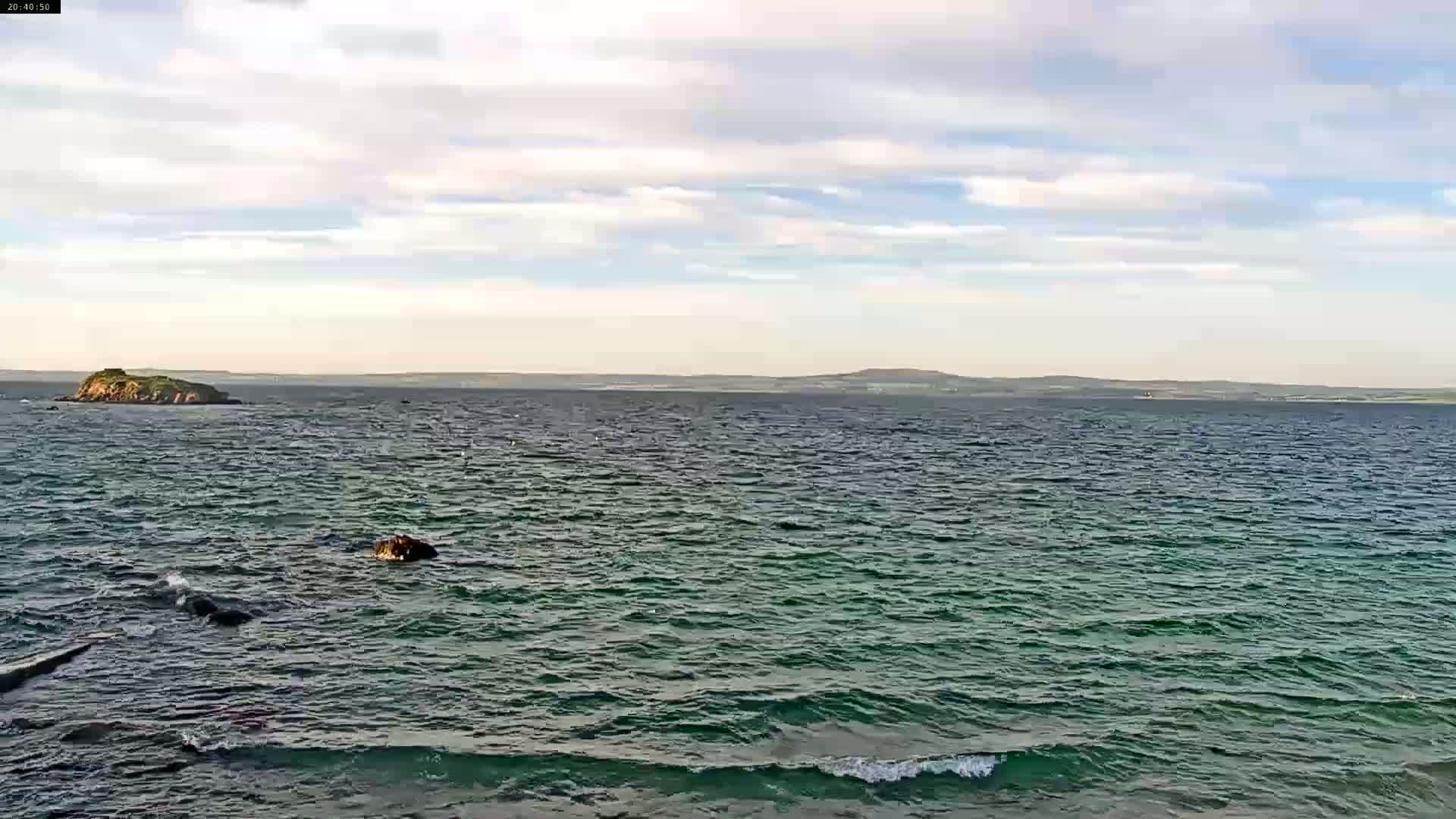 A partly cloudy day shows a body of water with small waves, a small island in the foreground, and a larger landmass in the distance.