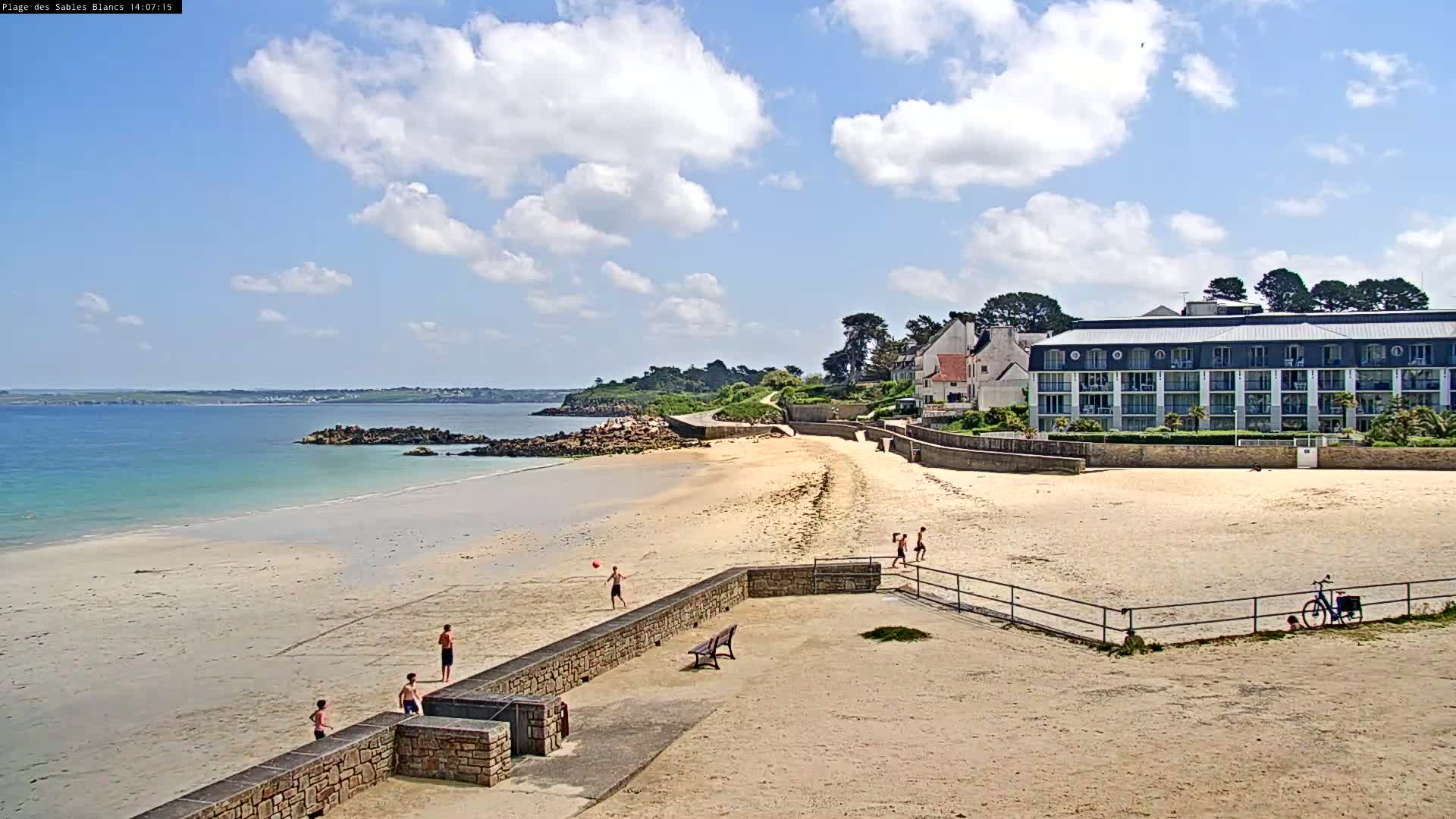 A sandy beach with several people enjoying the sunny day, a calm ocean, and a multi-story building nearby.