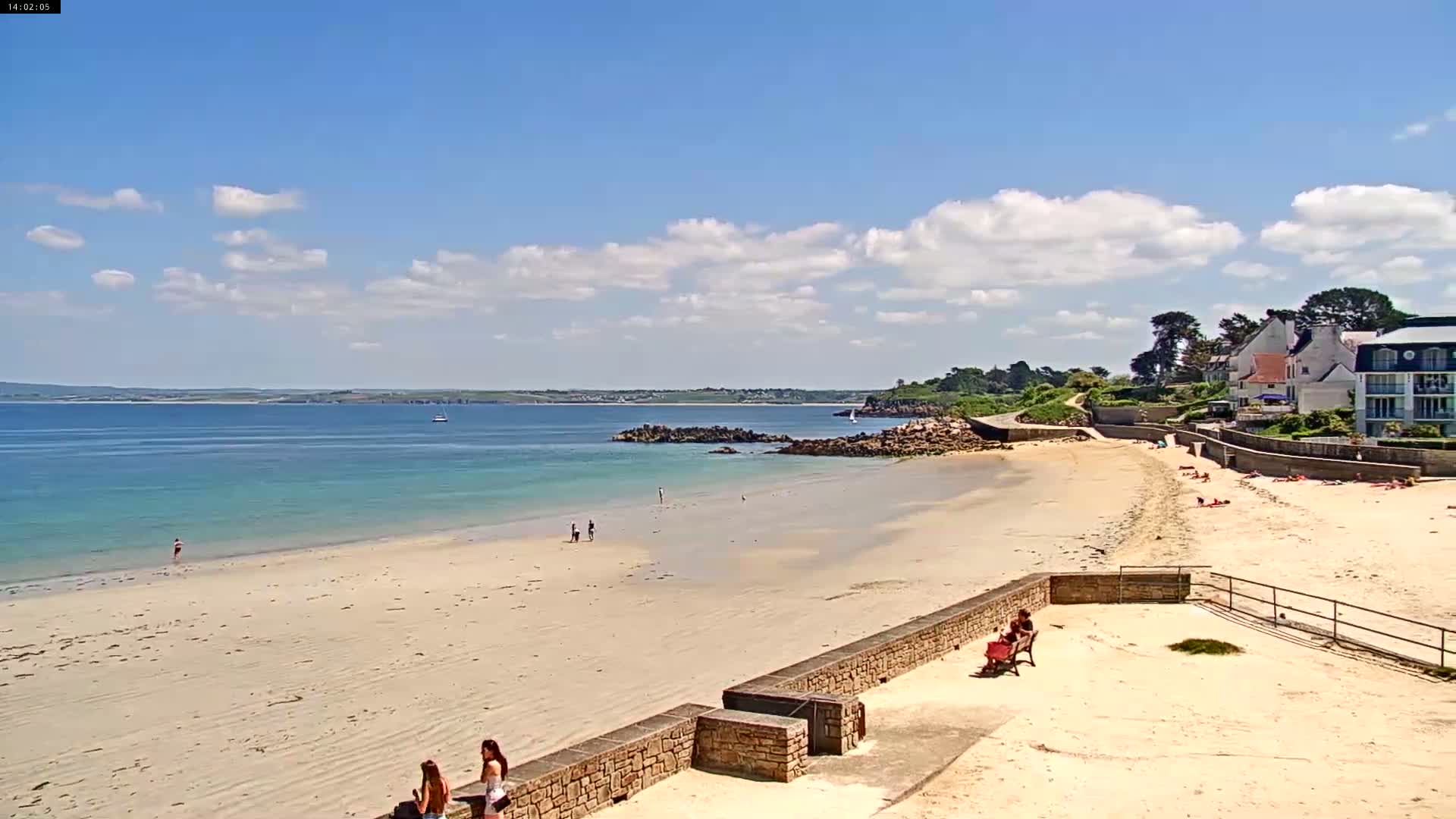 A sandy beach stretches out to calm, light-blue water under a mostly sunny sky with scattered clouds, and a few people are visible on the beach and in the water near a low stone wall and buildings.