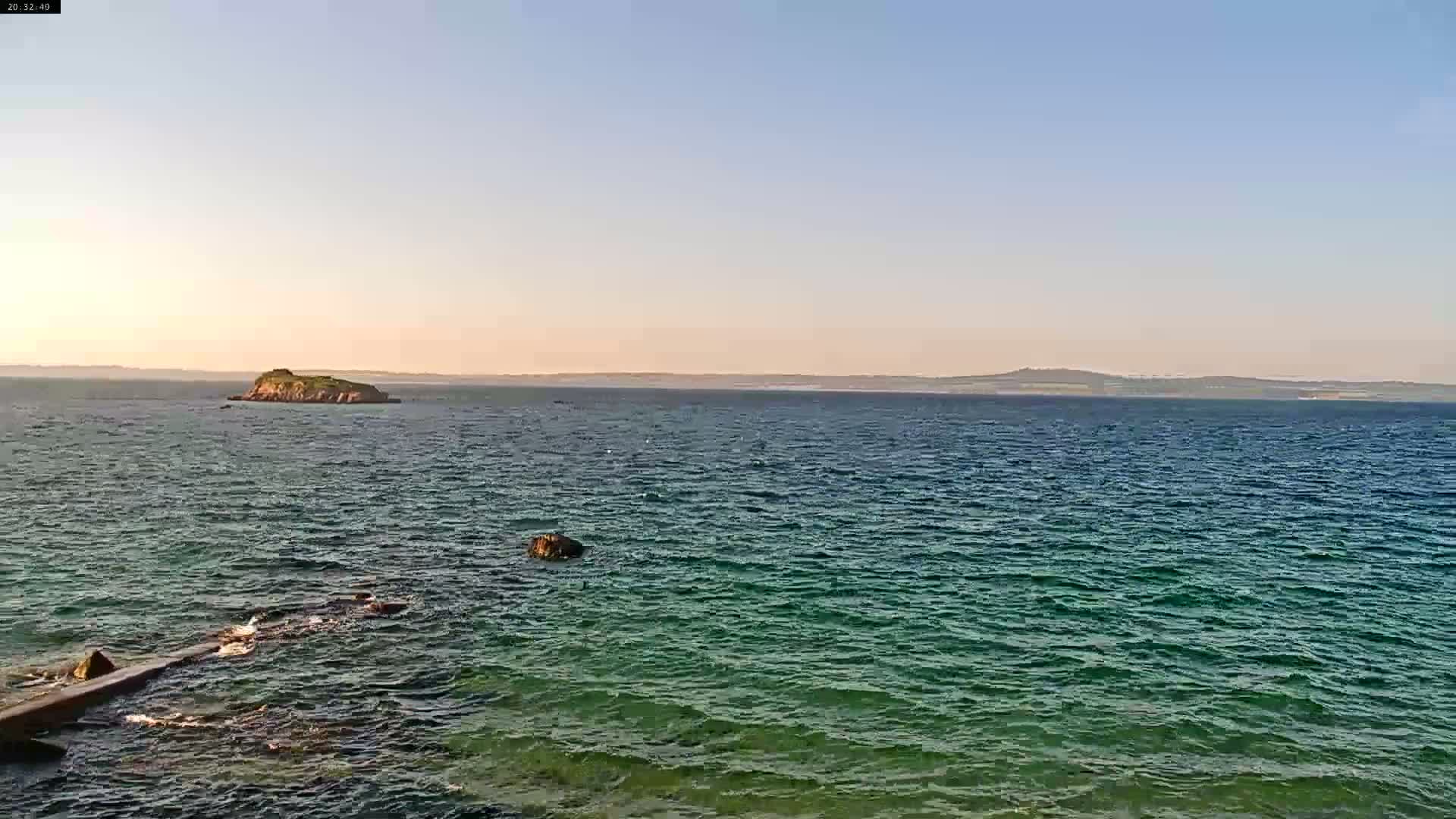 A calm sea with a small island in the mid-ground and a larger landmass on the horizon under a clear, light-blue sky.