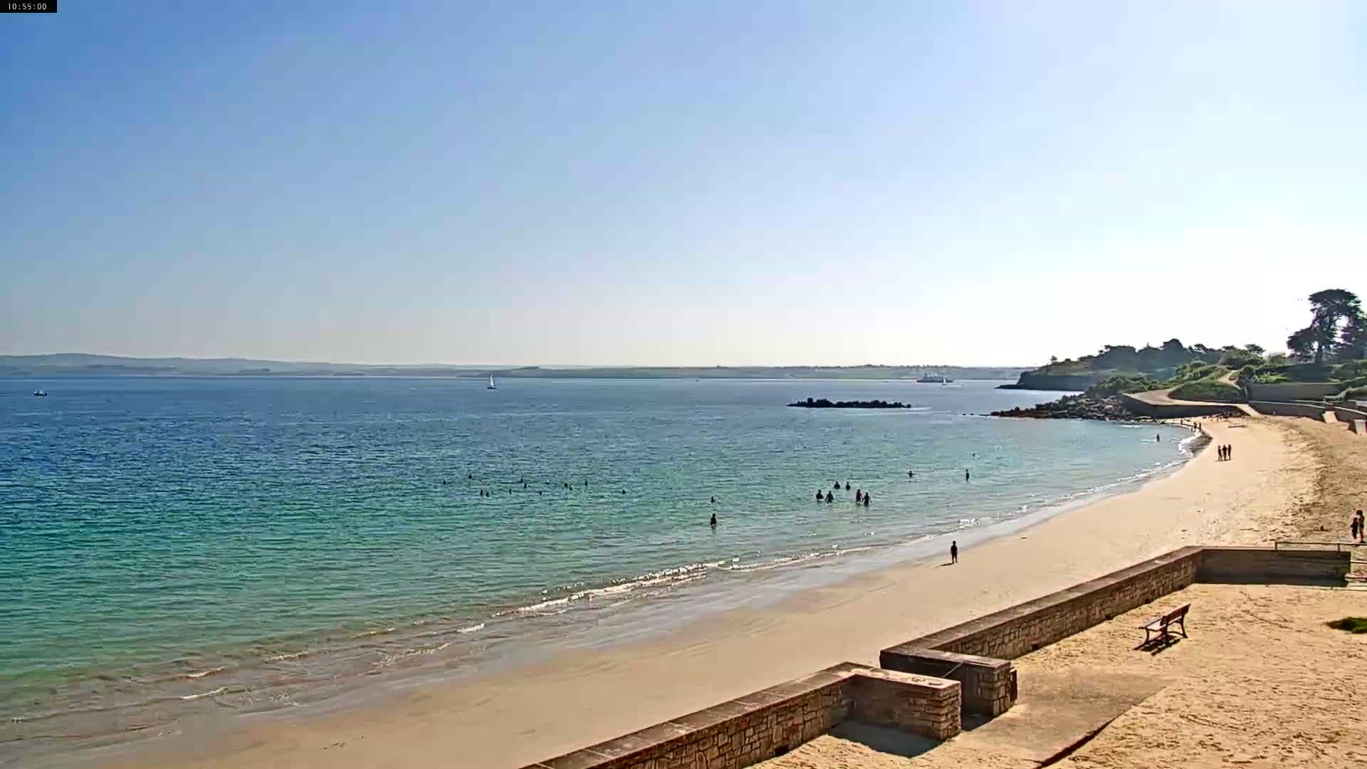 A sandy beach with calm, teal water stretches out to a clear blue sky, with a few people swimming and walking along the shore.