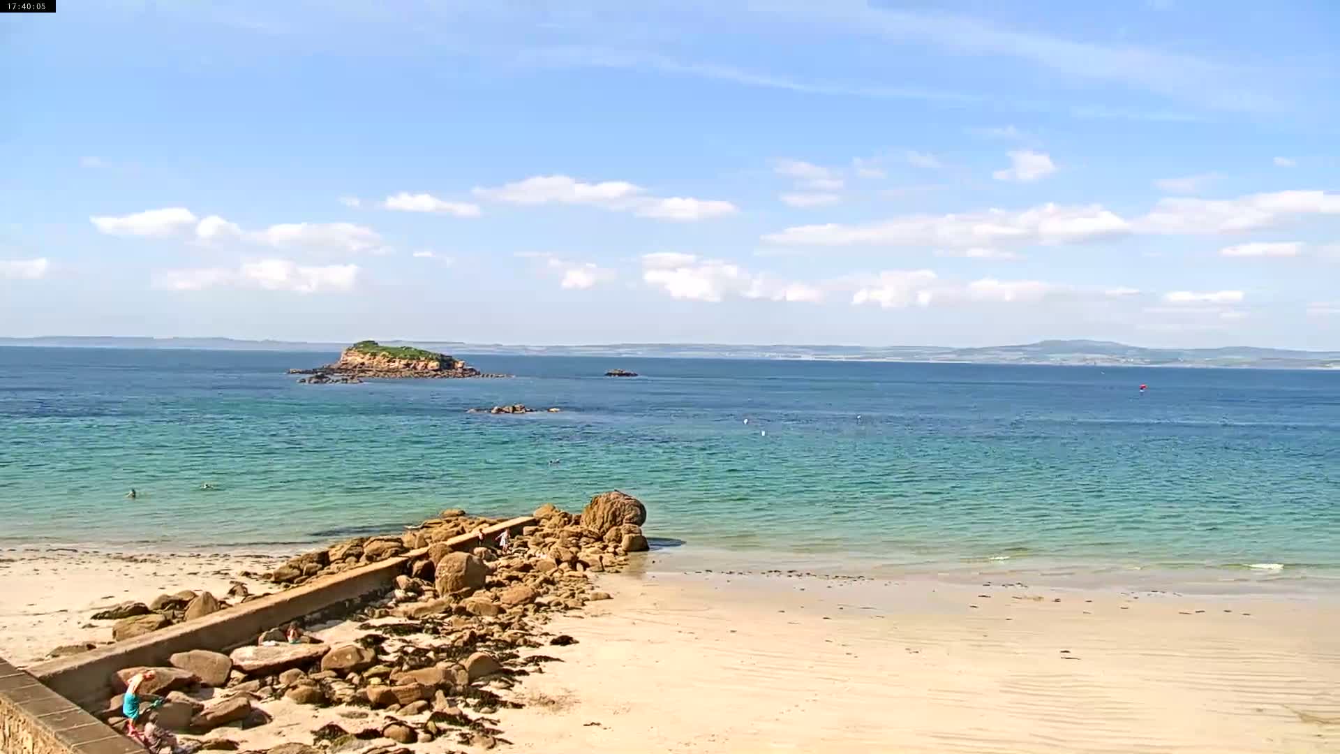 A sandy beach with a rocky shoreline and a calm, clear turquoise sea stretches to a distant island under a partly cloudy blue sky.