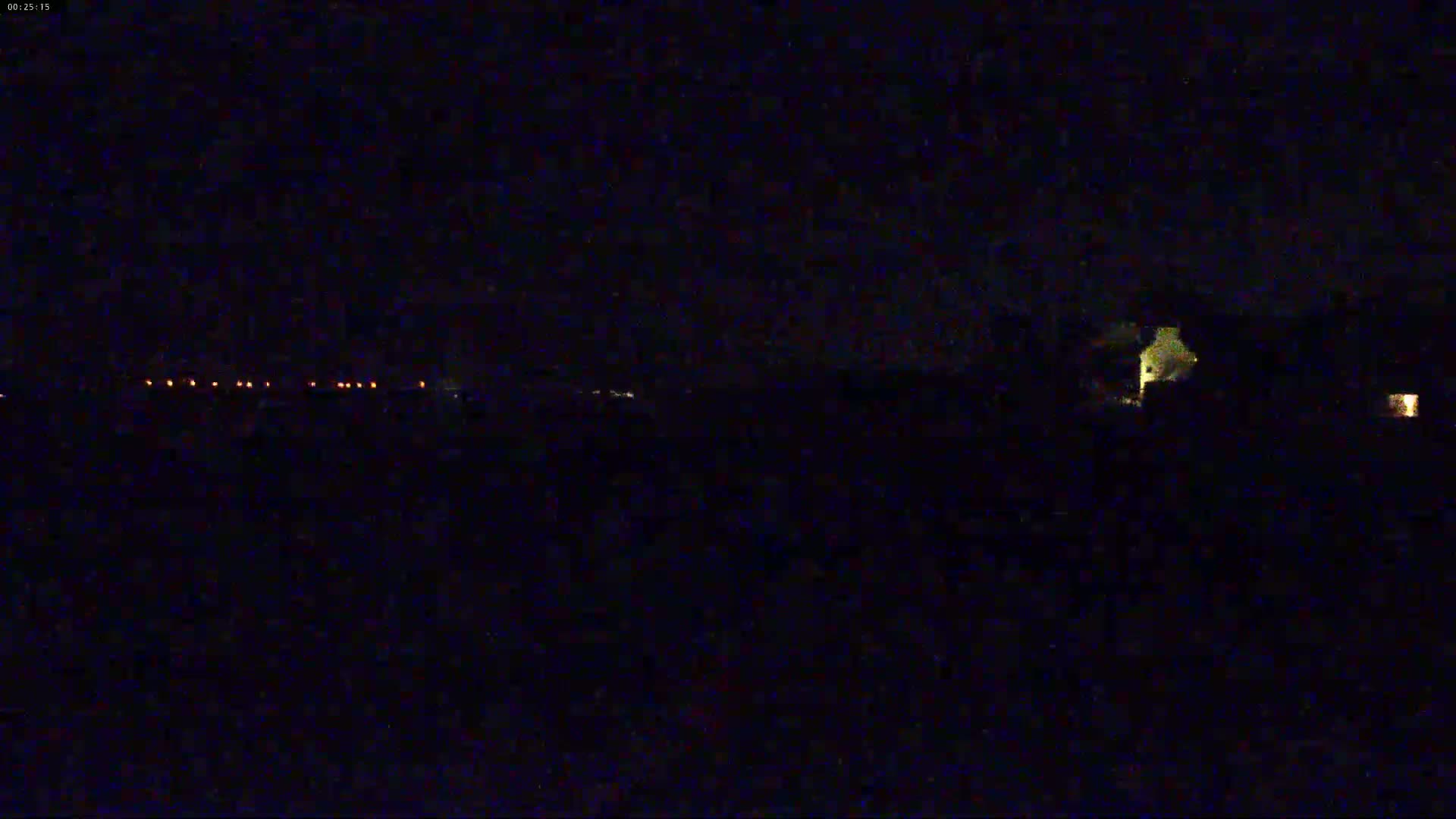 A nighttime, dark scene shows a line of distant lights and a faintly illuminated building in the foreground under a clear, dark sky.