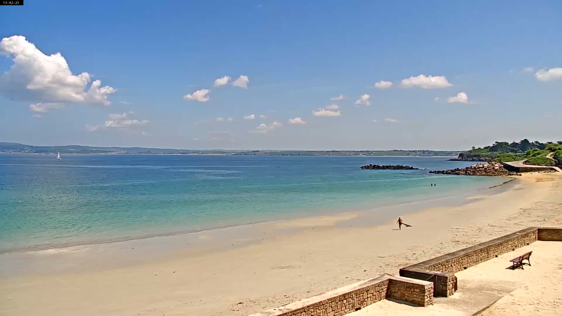 A sandy beach with calm, turquoise water stretches beneath a mostly sunny sky with scattered cumulus clouds, a lone sailboat visible in the distance.