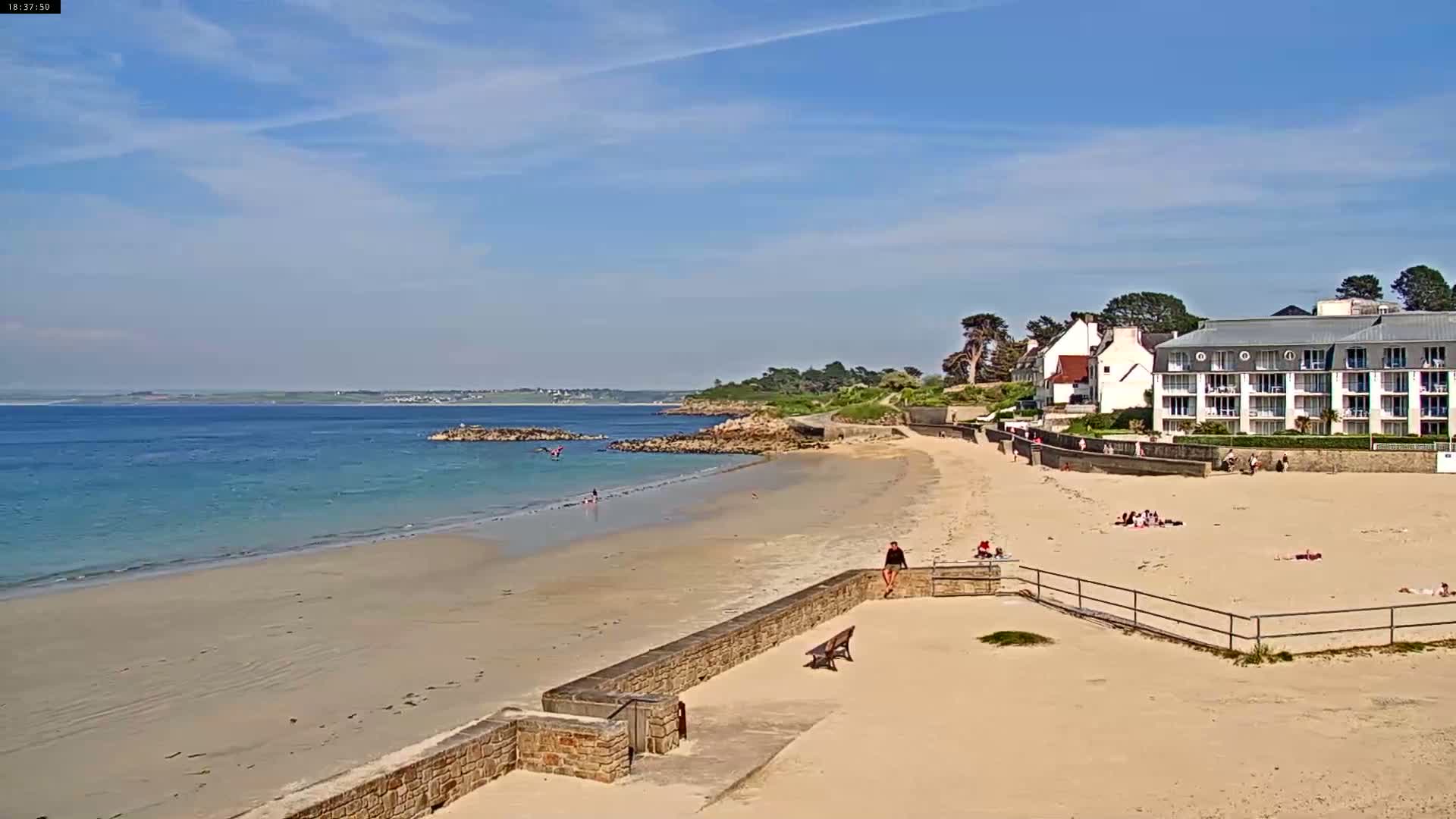 A sandy beach with calm, blue water stretches out to a distant coastline under a mostly sunny sky, with a few people relaxing on the sand and near a low stone wall separating the beach from a building.