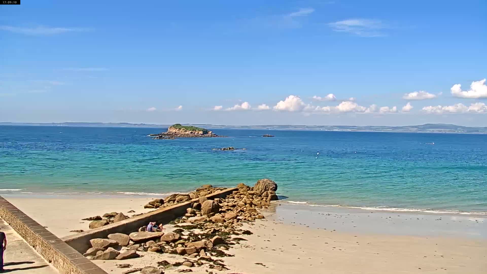 A sandy beach with a rocky outcrop and a small island in the distance under a mostly sunny sky.