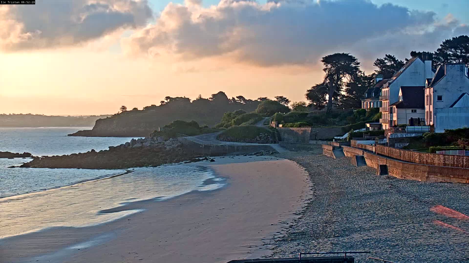 A sandy beach curves gently towards a calm ocean under a partly cloudy sunset sky, with several houses situated on a hillside overlooking the scene.