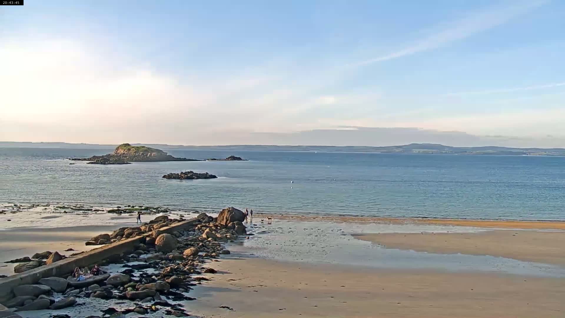A sandy beach with a rocky breakwater stretches toward a calm, blue ocean with a small island in the distance under a mostly clear sky.