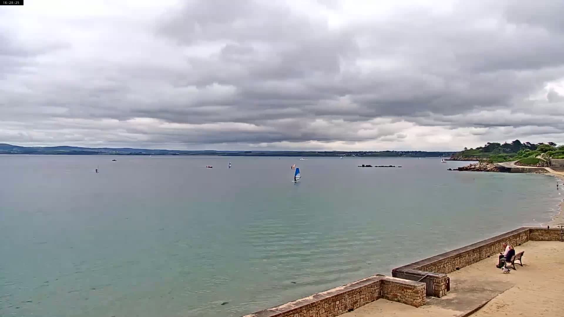 A calm, overcast day shows a view of a bay with several sailboats and windsurfers, a sandy beach, and a stone wall with people sitting on a bench.
