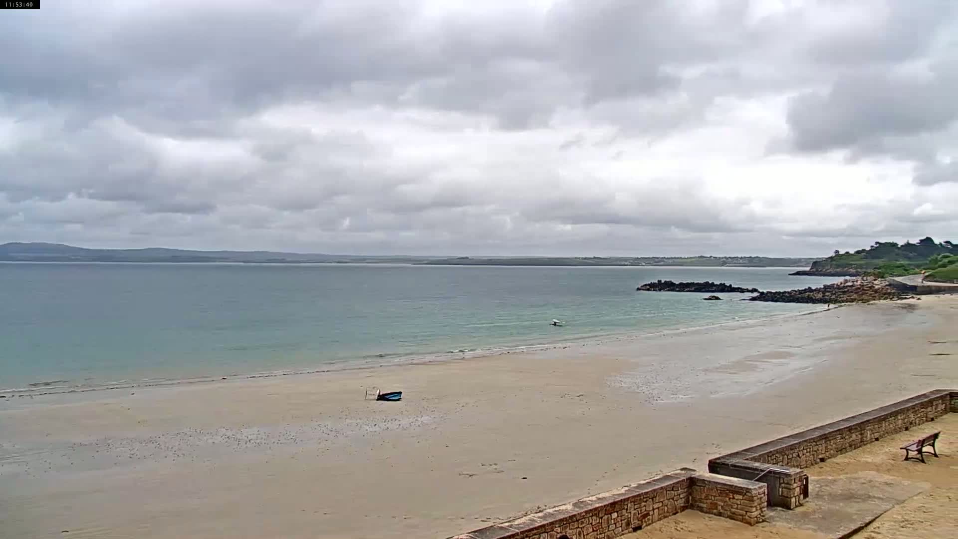 A sandy beach stretches out to a calm sea under a cloudy sky, with a small boat resting on the sand and a rocky outcrop visible in the distance.