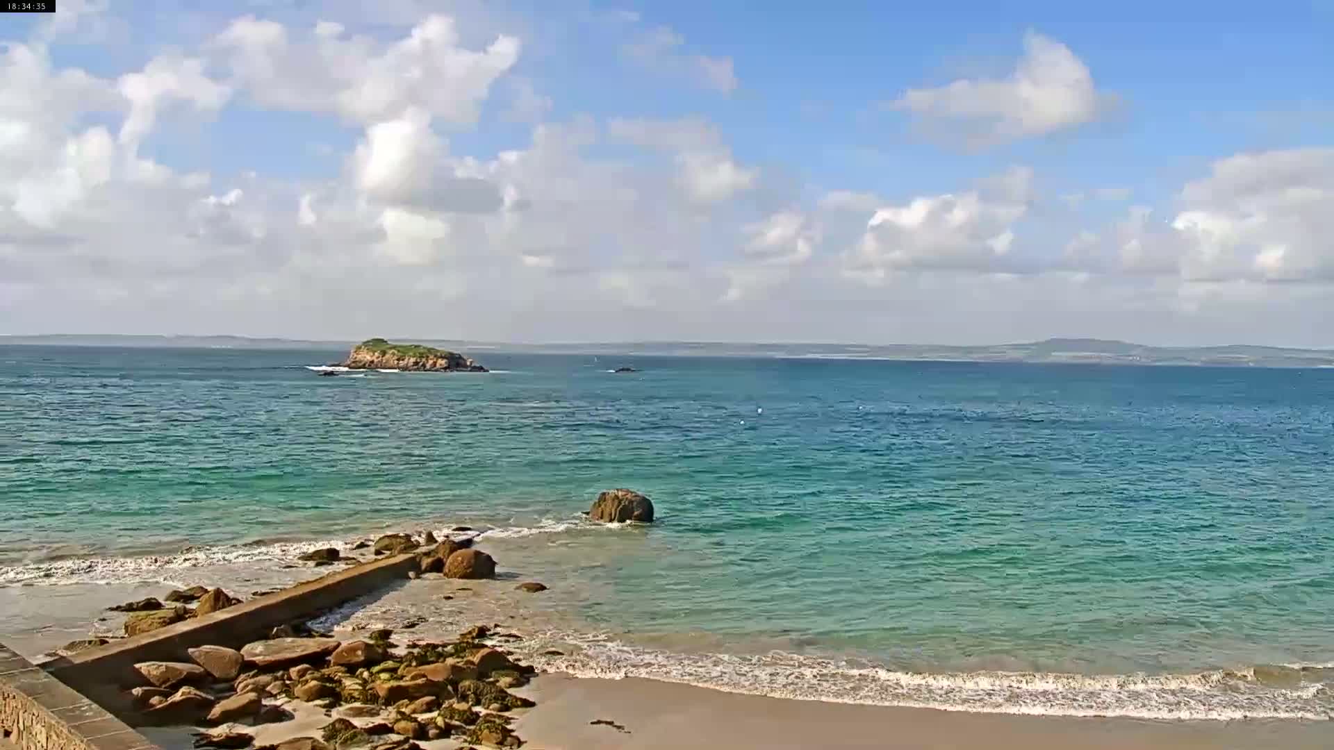 A partly cloudy day shows a tranquil ocean scene with a small island in the distance, gentle waves lapping a sandy beach with rocks and a low wall.