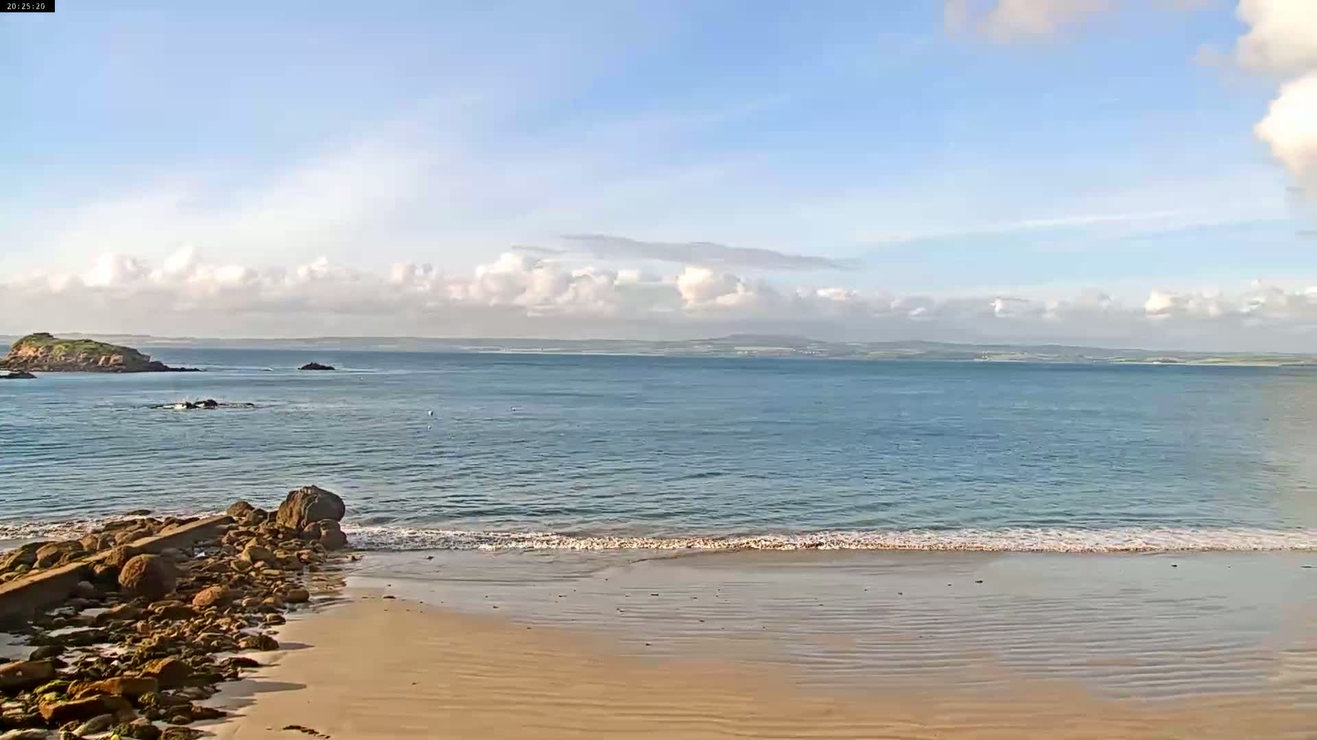 A sandy beach with rocky outcroppings meets a calm sea under a partly cloudy sky.