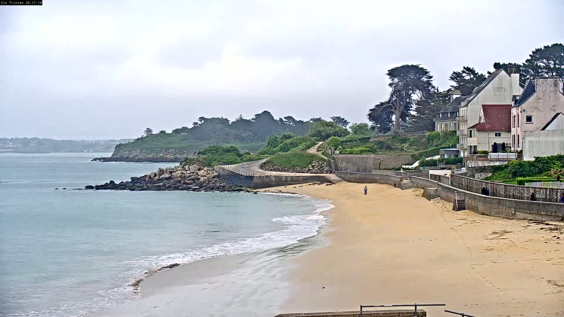A sandy beach curves gently toward a calm, light-blue ocean under an overcast sky, bordered by a rocky shoreline and a row of houses perched on a verdant hillside.