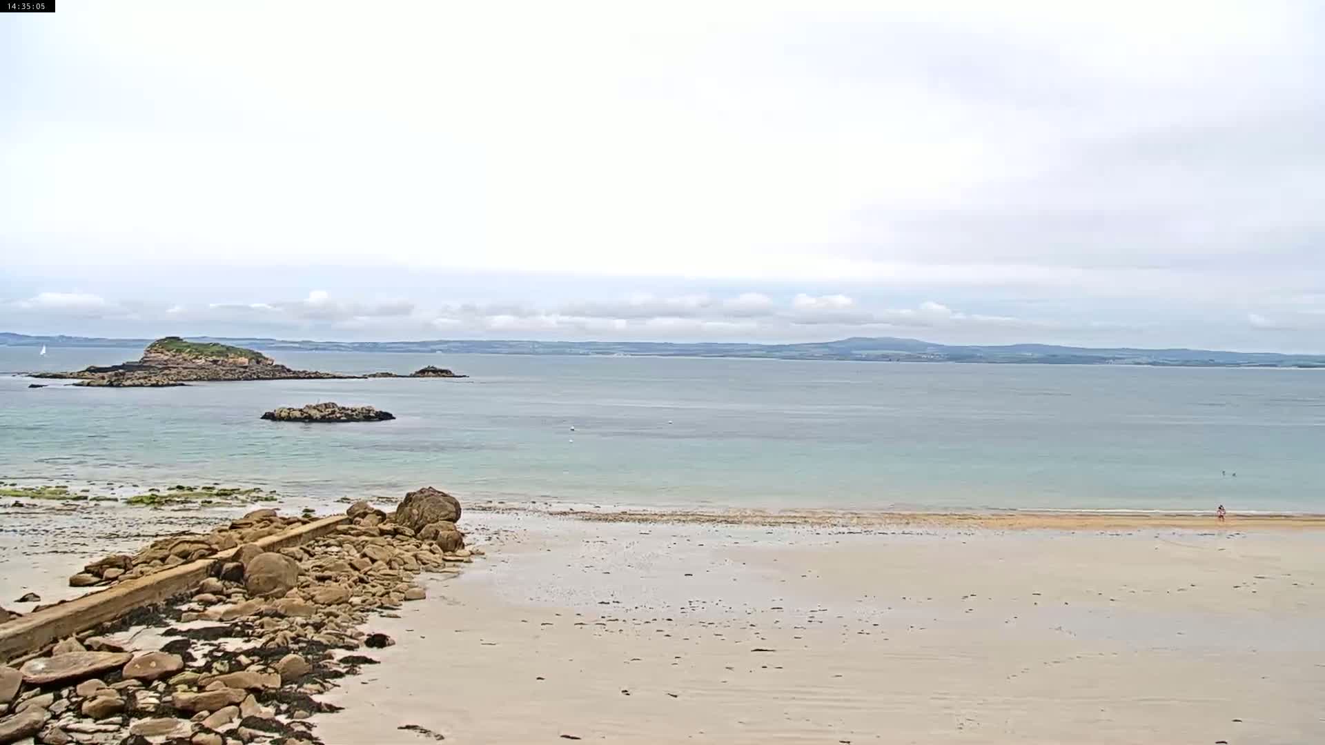 A sandy beach with rocky outcroppings and a calm, light-blue ocean under a cloudy sky stretches to a distant coastline.