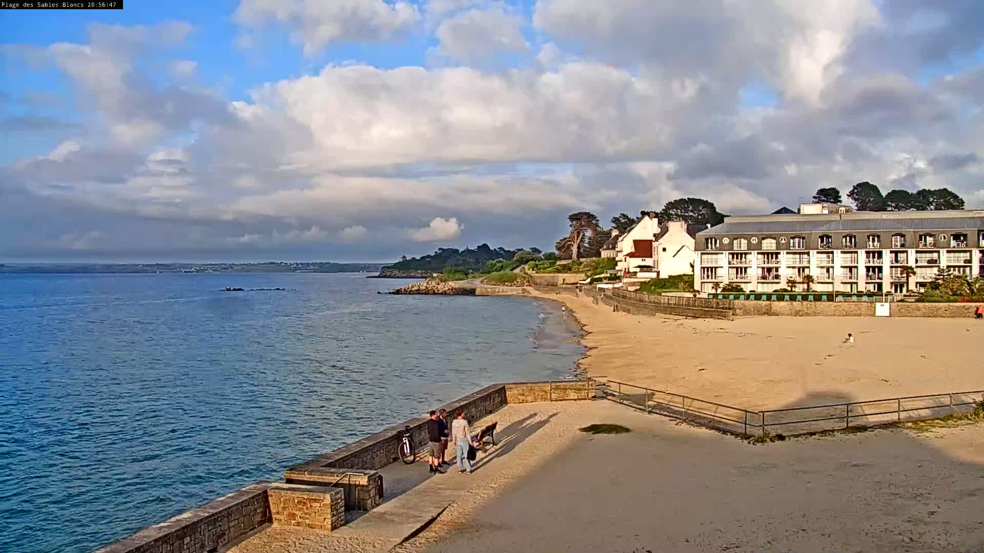 A sandy beach with a multi-story building sits beside calm blue water under a partly cloudy sky.