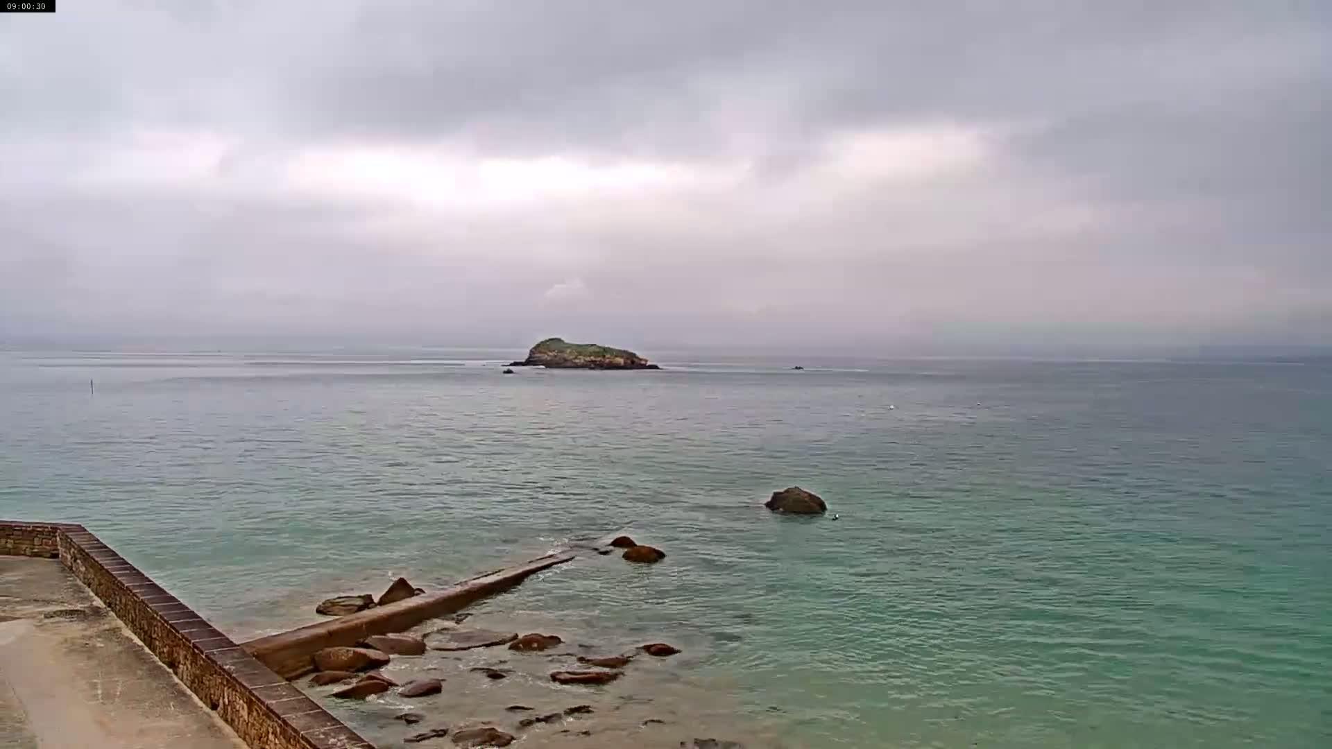 A calm, overcast day shows a rocky shoreline with a small island in the distance.