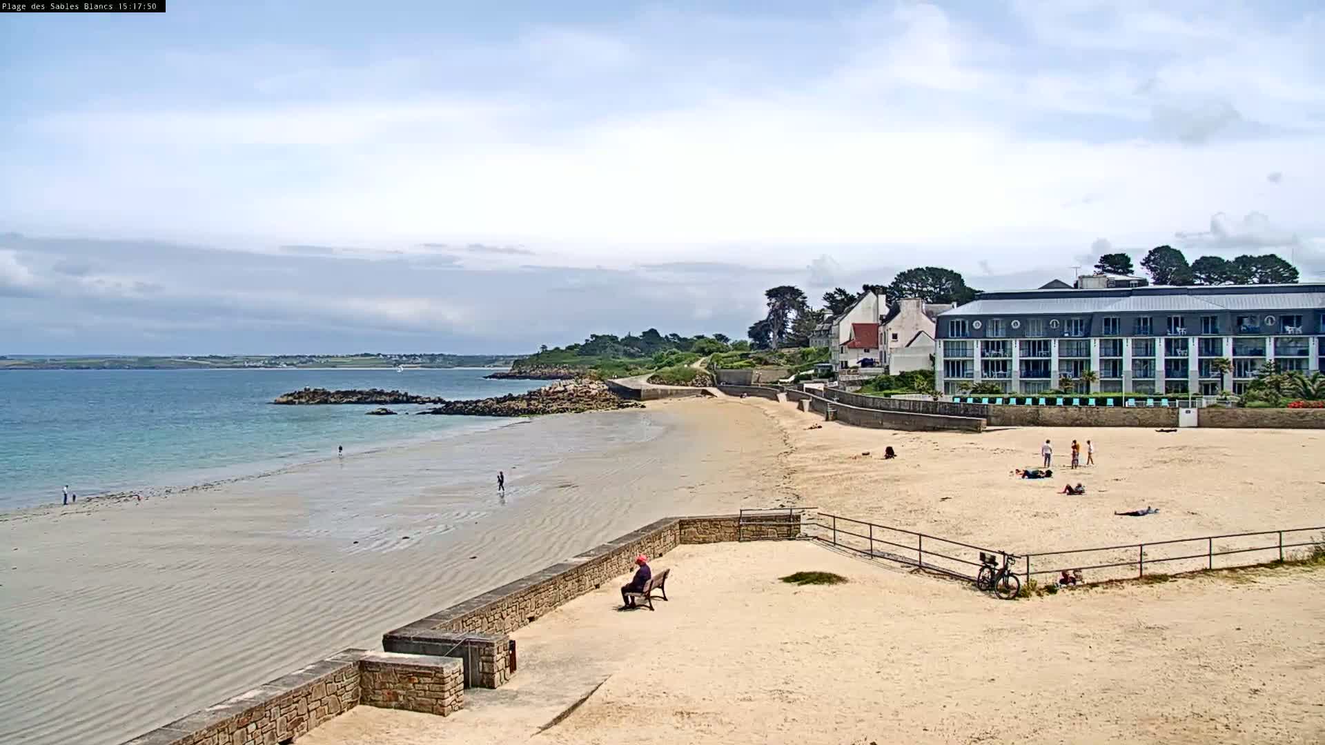 A sandy beach stretches along a calm ocean under a partly cloudy sky, with a multi-story building and several people visible on the sand.