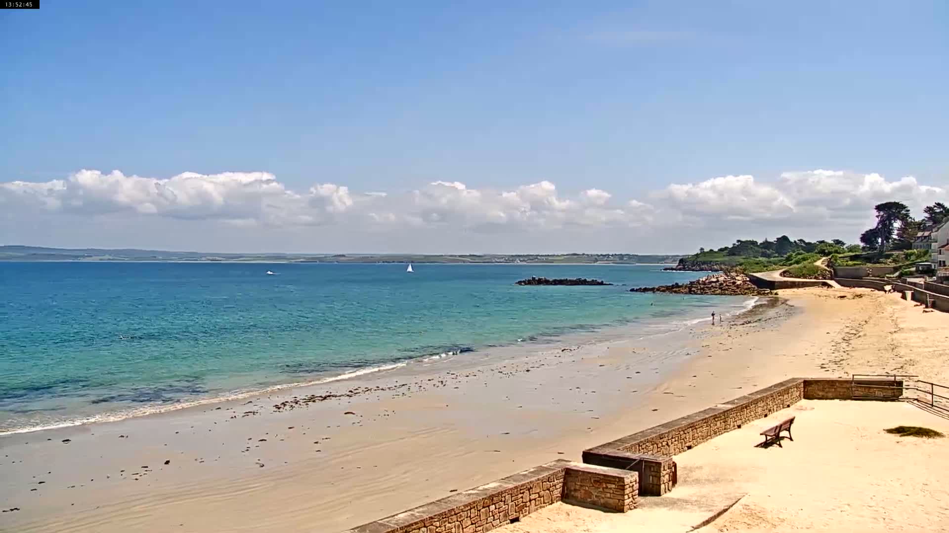 A sandy beach with calm, light-blue water stretches under a partly cloudy, sunny sky.