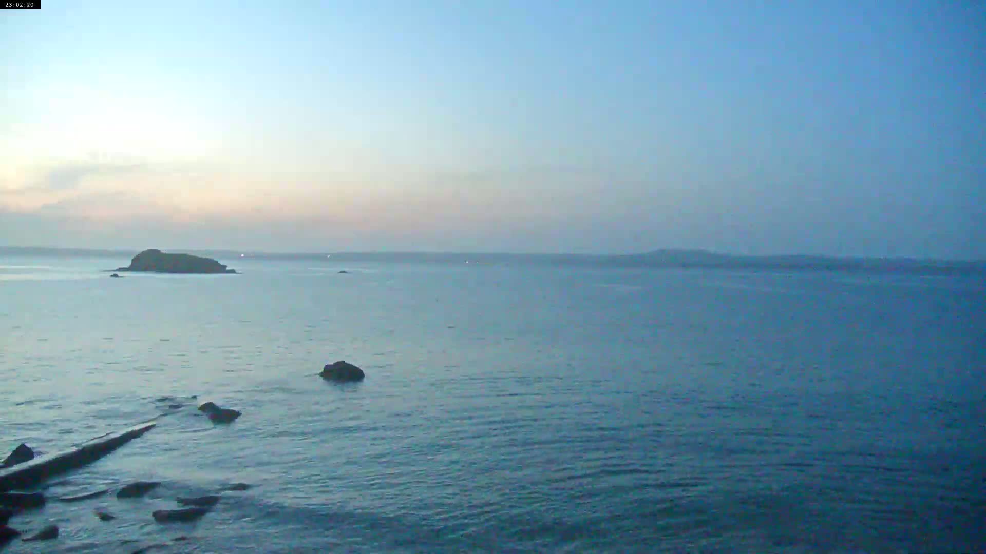 A calm, twilight ocean scene shows a small island in the mid-ground and a rocky shoreline in the foreground.
