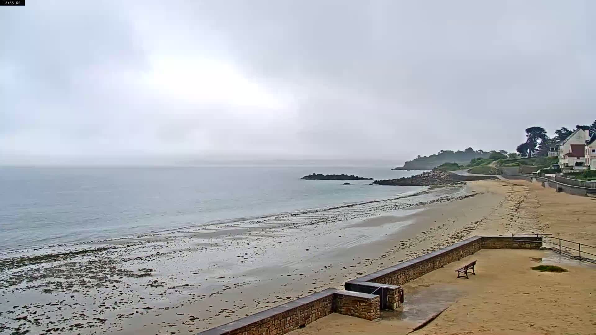 A sandy beach stretches out to a calm, gray sea under an overcast sky, with a stone wall and a few buildings visible in the distance.