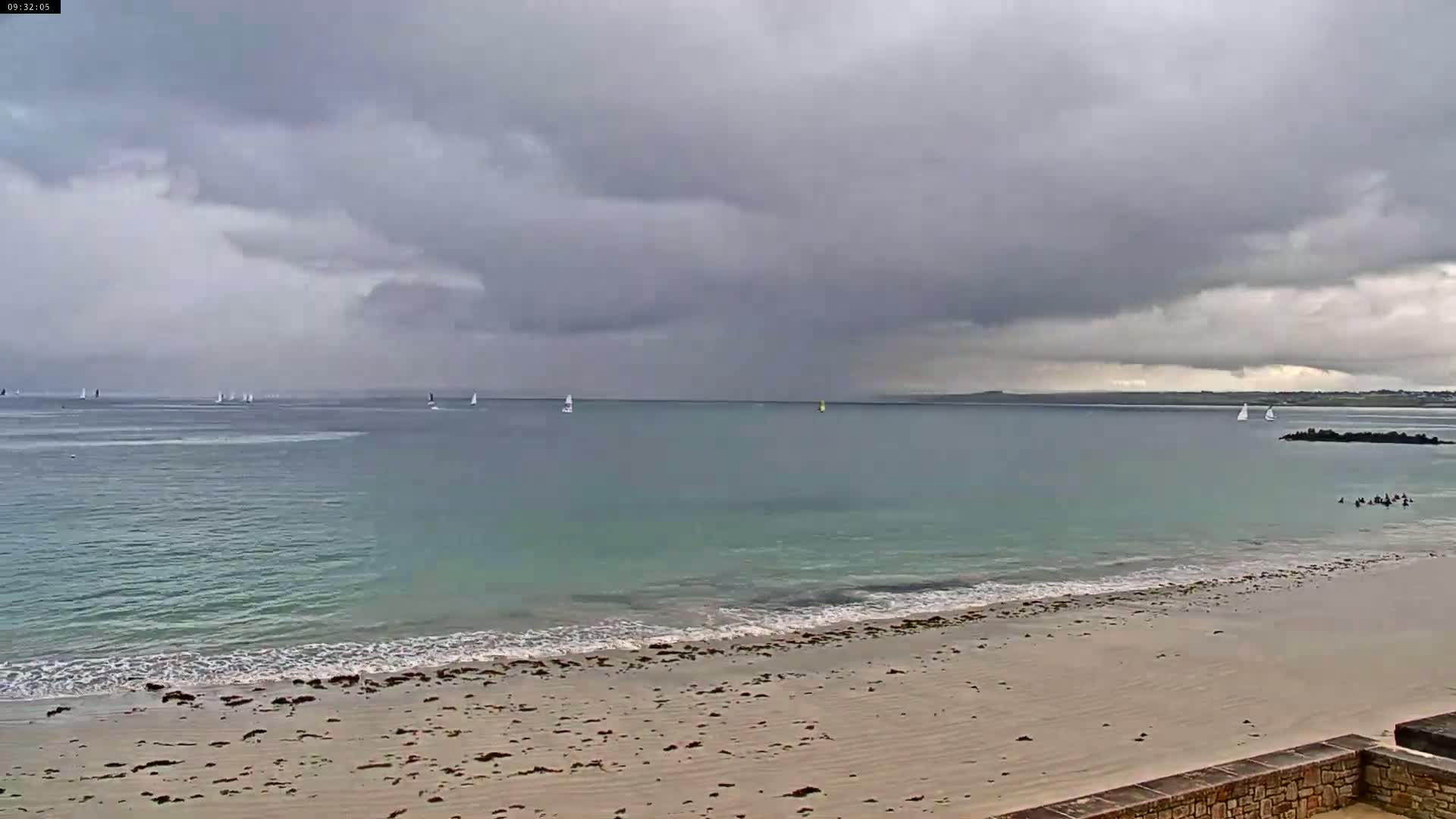 A sandy beach with a calm, light-green ocean under a heavily overcast sky shows several sailboats in the distance.