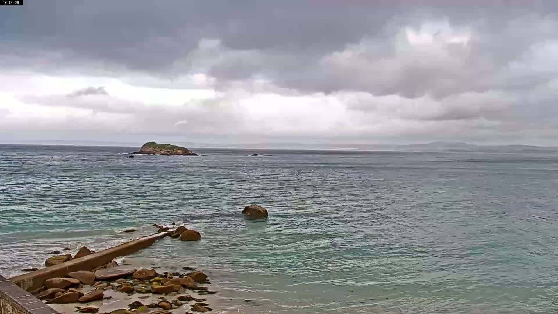 A rocky shoreline with a low wall extends into a calm, teal ocean under a mostly cloudy, gray sky, with a small island visible in the distance.