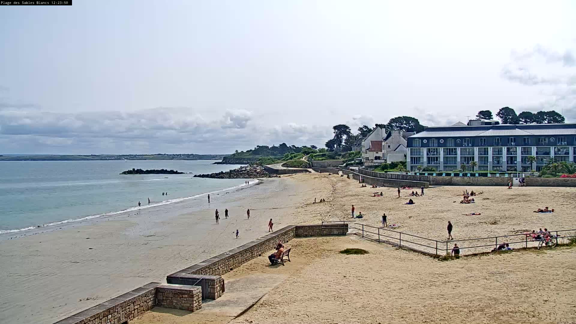 A sandy beach with scattered sunbathers is situated next to a calm ocean and a large building under a partly cloudy sky.