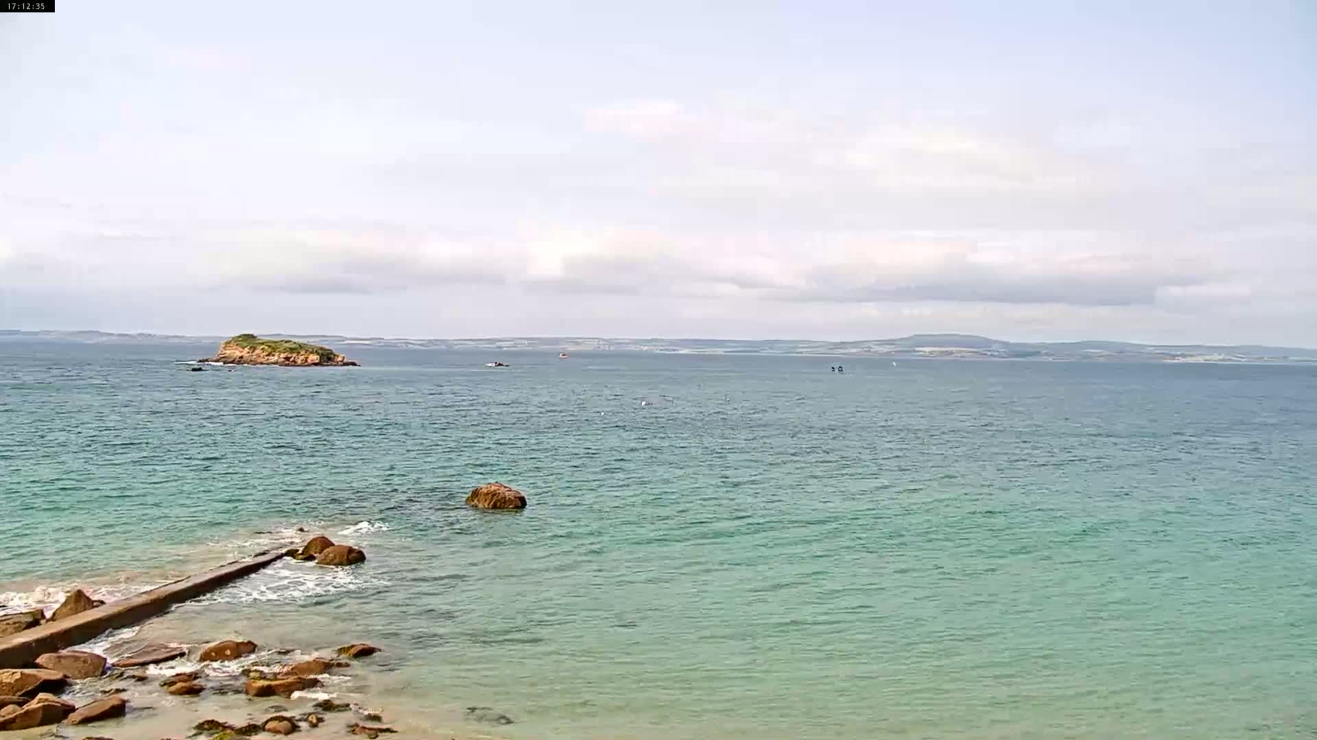 A rocky shoreline meets a calm, teal ocean under a mostly cloudy sky, with a small island visible in the distance.