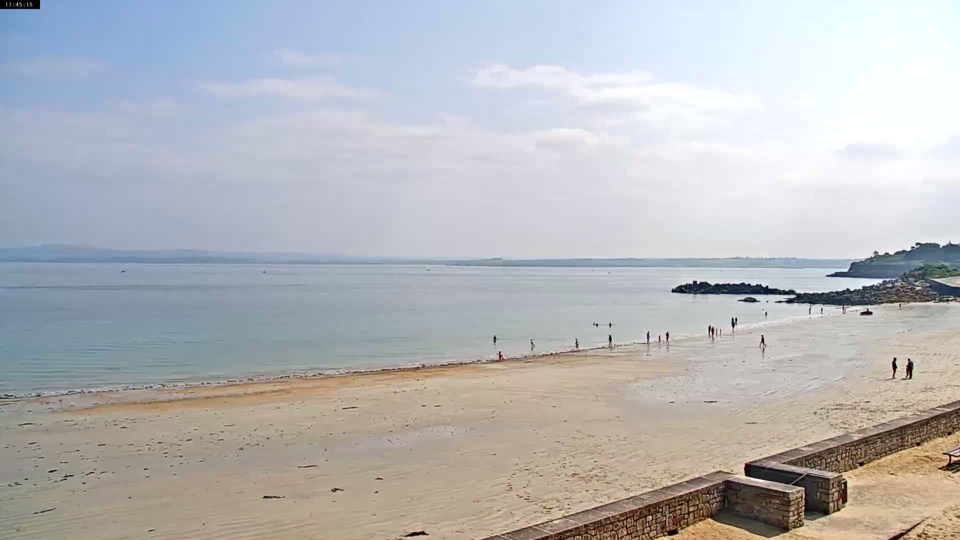 A mostly sunny day at a wide sandy beach with calm, light-blue water, a few people wading and walking along the shore, and a rocky outcrop at the right.