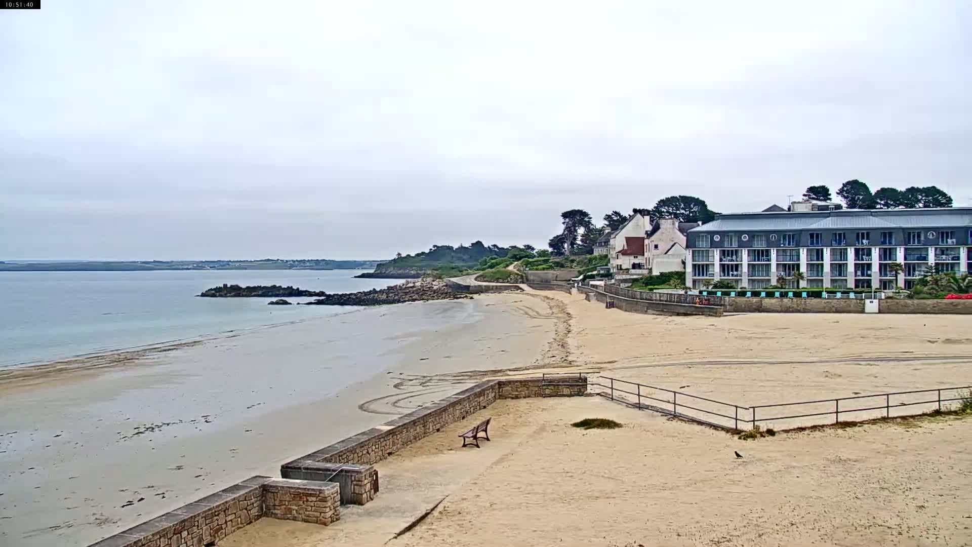 A sandy beach stretches out to a calm body of water under an overcast sky, with buildings and greenery visible along the shoreline.