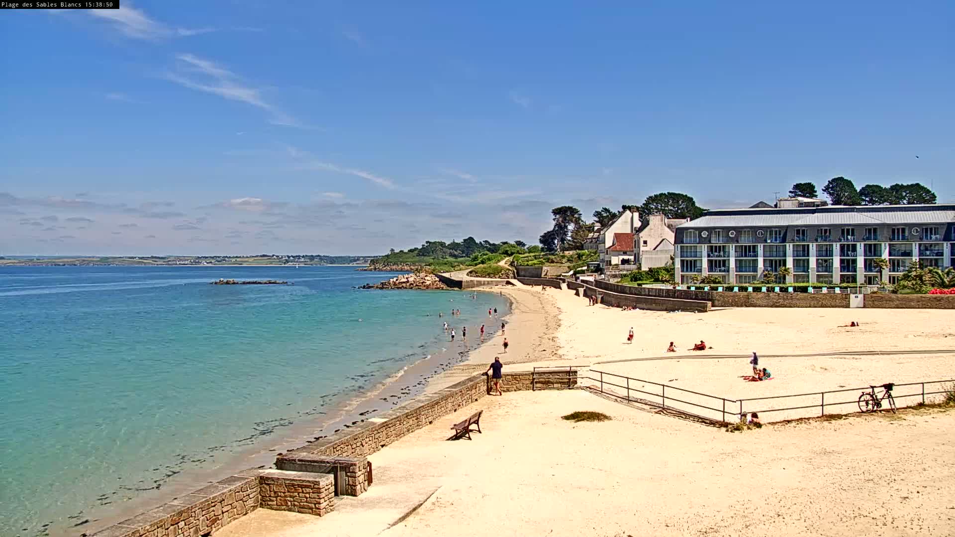 A sandy beach with calm, turquoise water stretches out beneath a mostly sunny sky, with a few people enjoying the beach and a large building visible beyond.