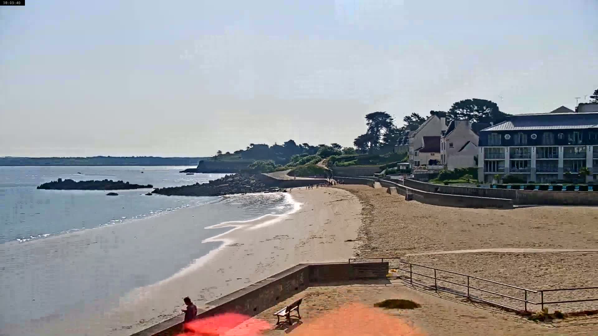 A sandy beach with calm water stretches out before a low stone wall, behind which are buildings and green vegetation, all under a clear, sunny sky.