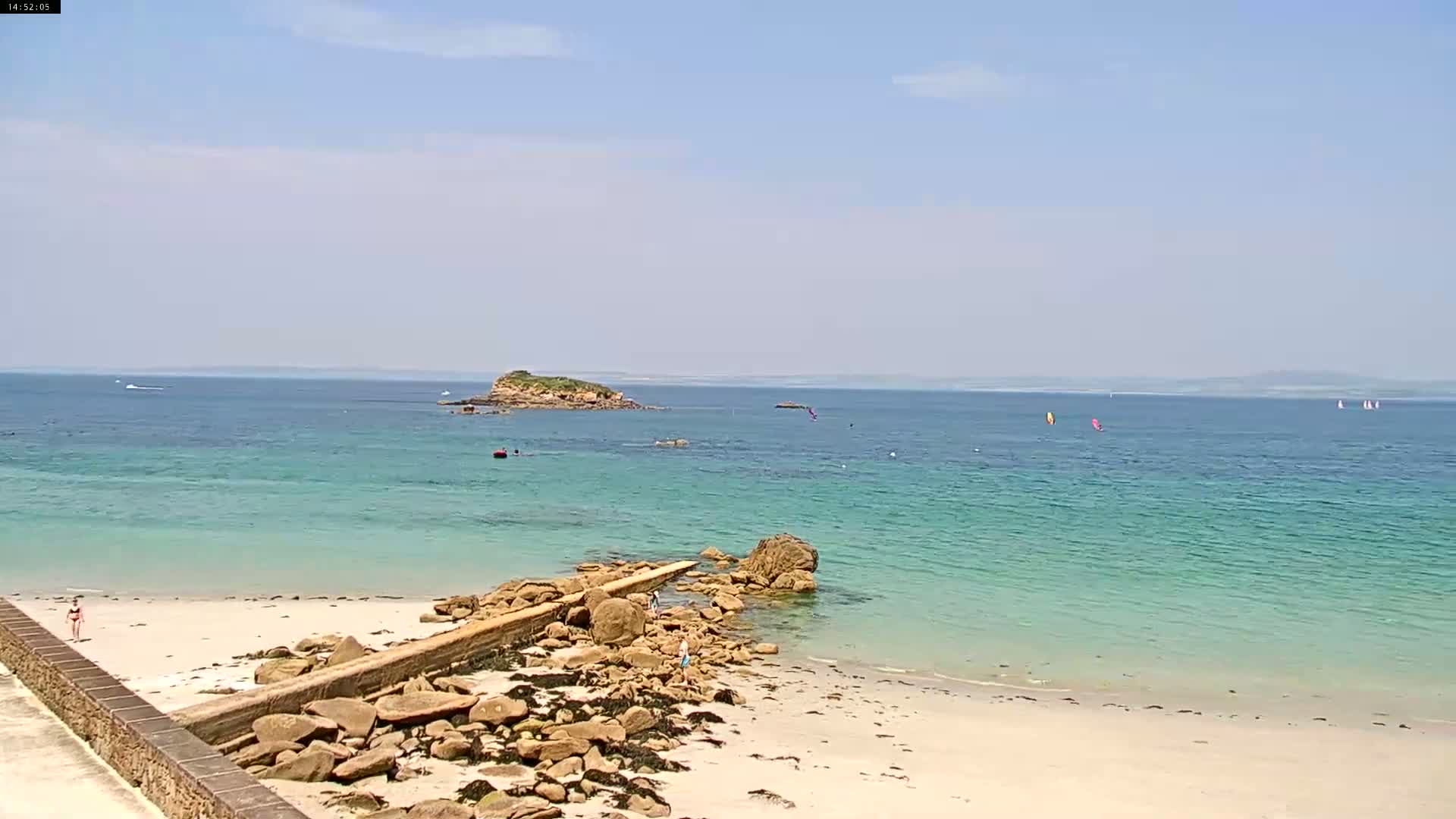 A sandy beach with clear, teal water meets a rocky shoreline under a mostly sunny sky; small boats and windsurfers are visible in the distance.