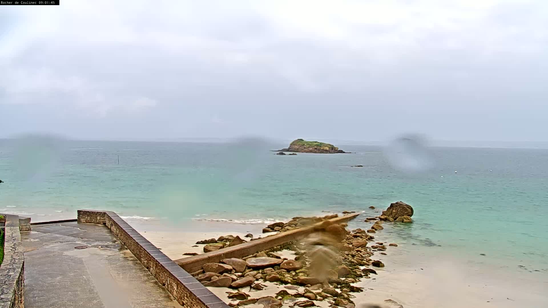 A rocky shoreline with a low wall leads to a sandy beach and light turquoise water, where a small island is visible under an overcast sky during rainfall.