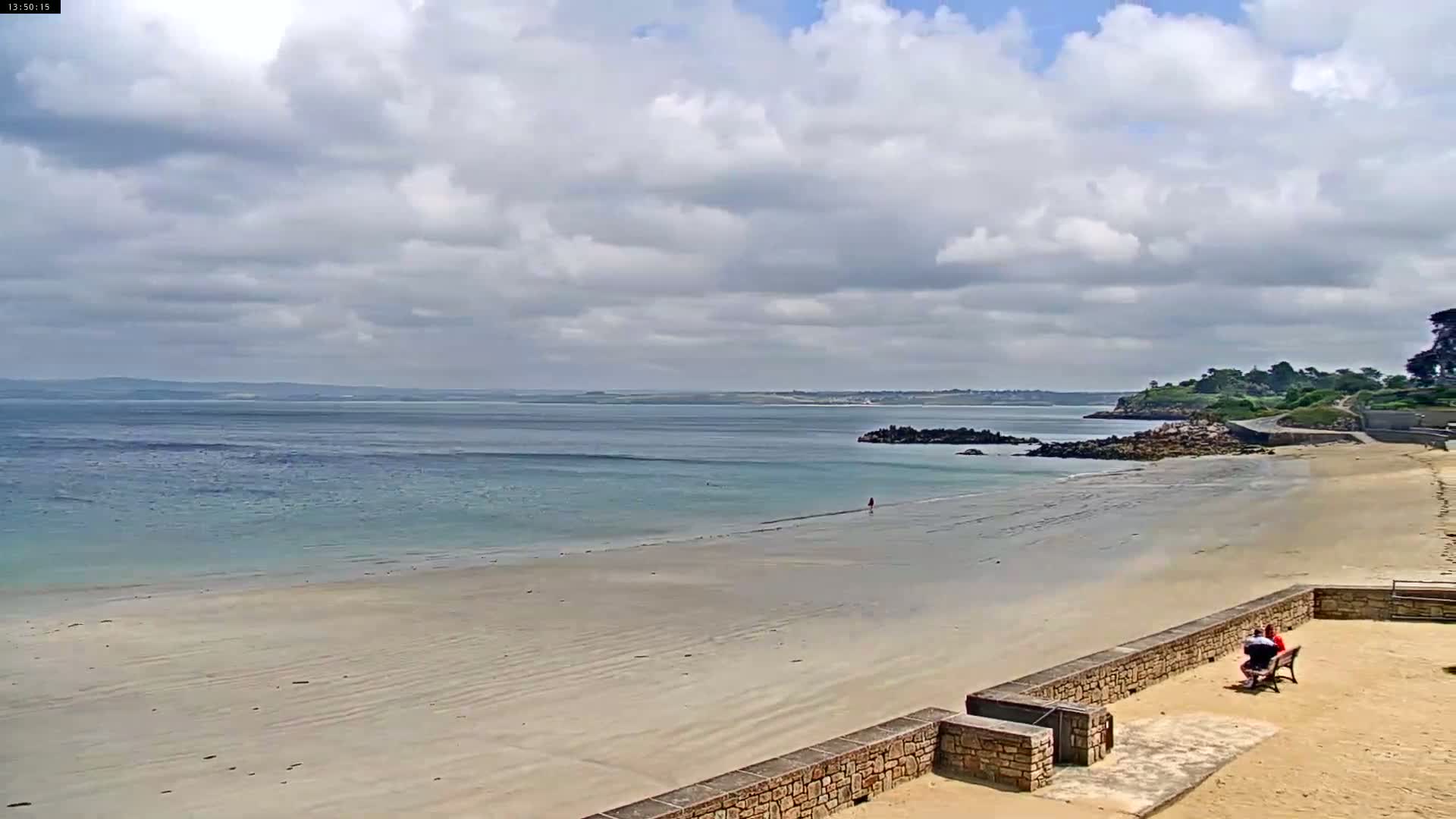 A sandy beach stretches out to a calm, light blue ocean under a partly cloudy sky, with a few people visible on the beach and a couple sitting on a bench.