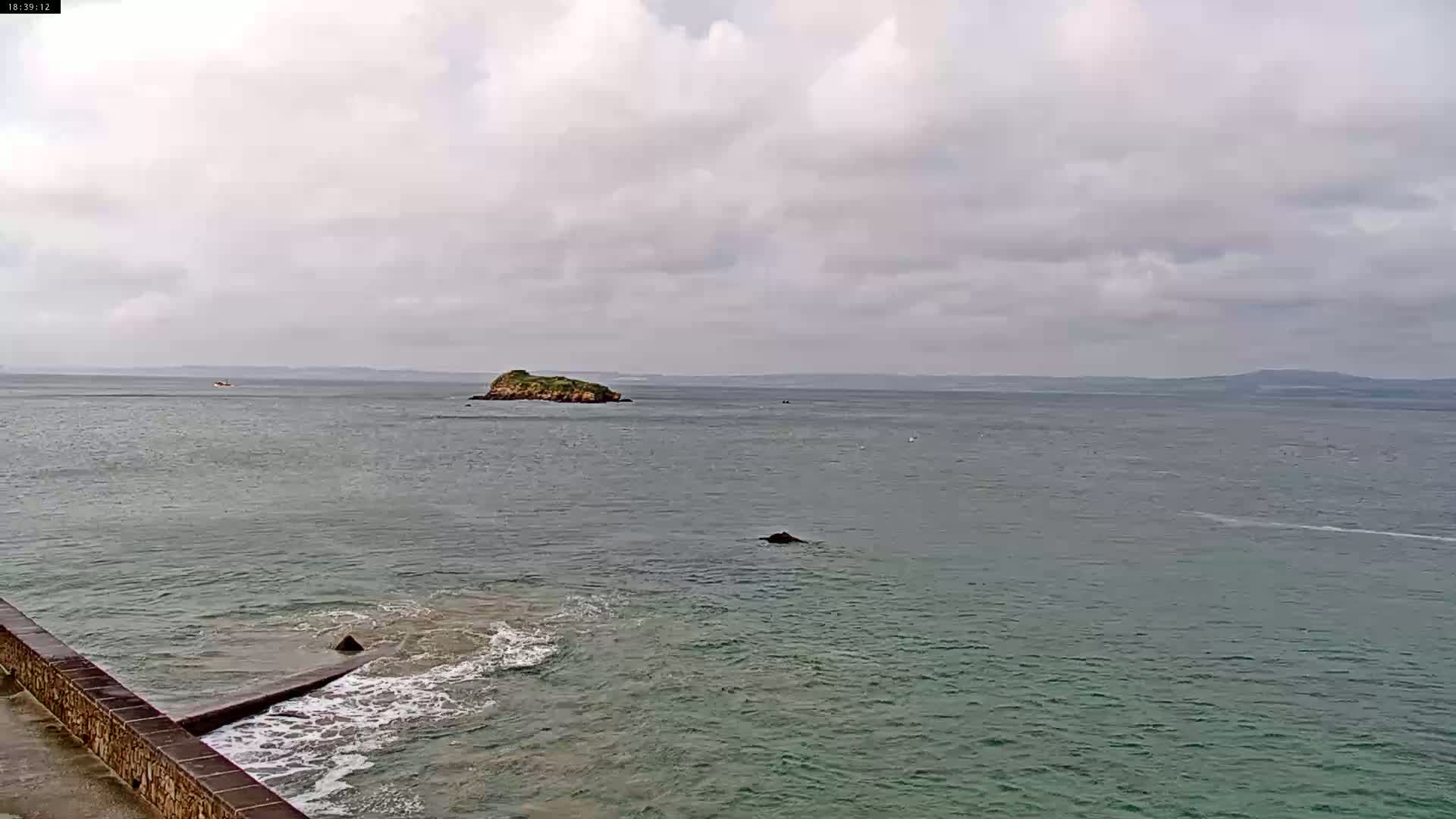 A calm, overcast day shows a view of a small island in a body of water, with a stone wall and small waves visible in the foreground.