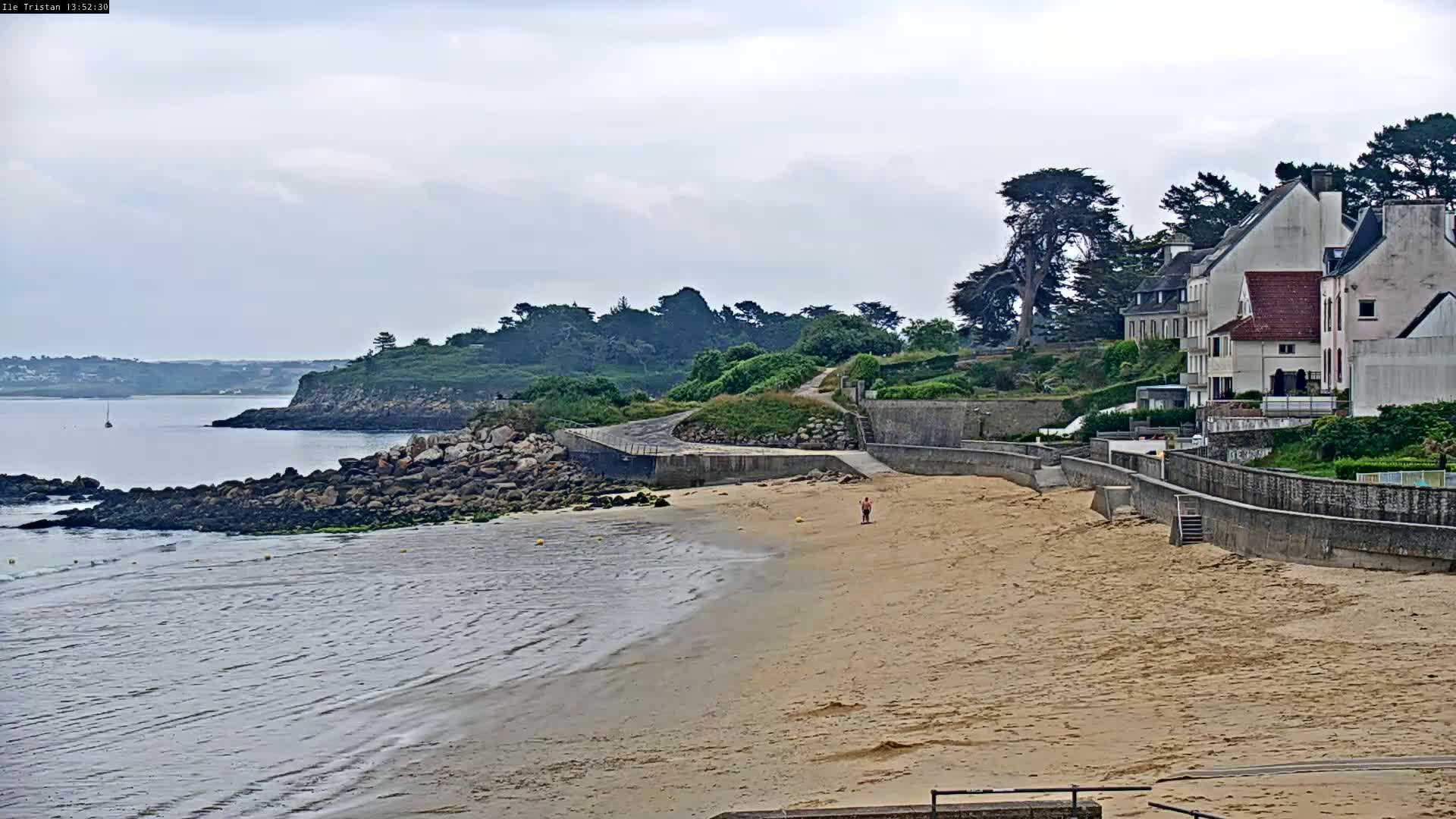 A sandy beach with calm, shallow water stretches out to a rocky shoreline, beyond which sits a small village of white buildings under a cloudy sky.