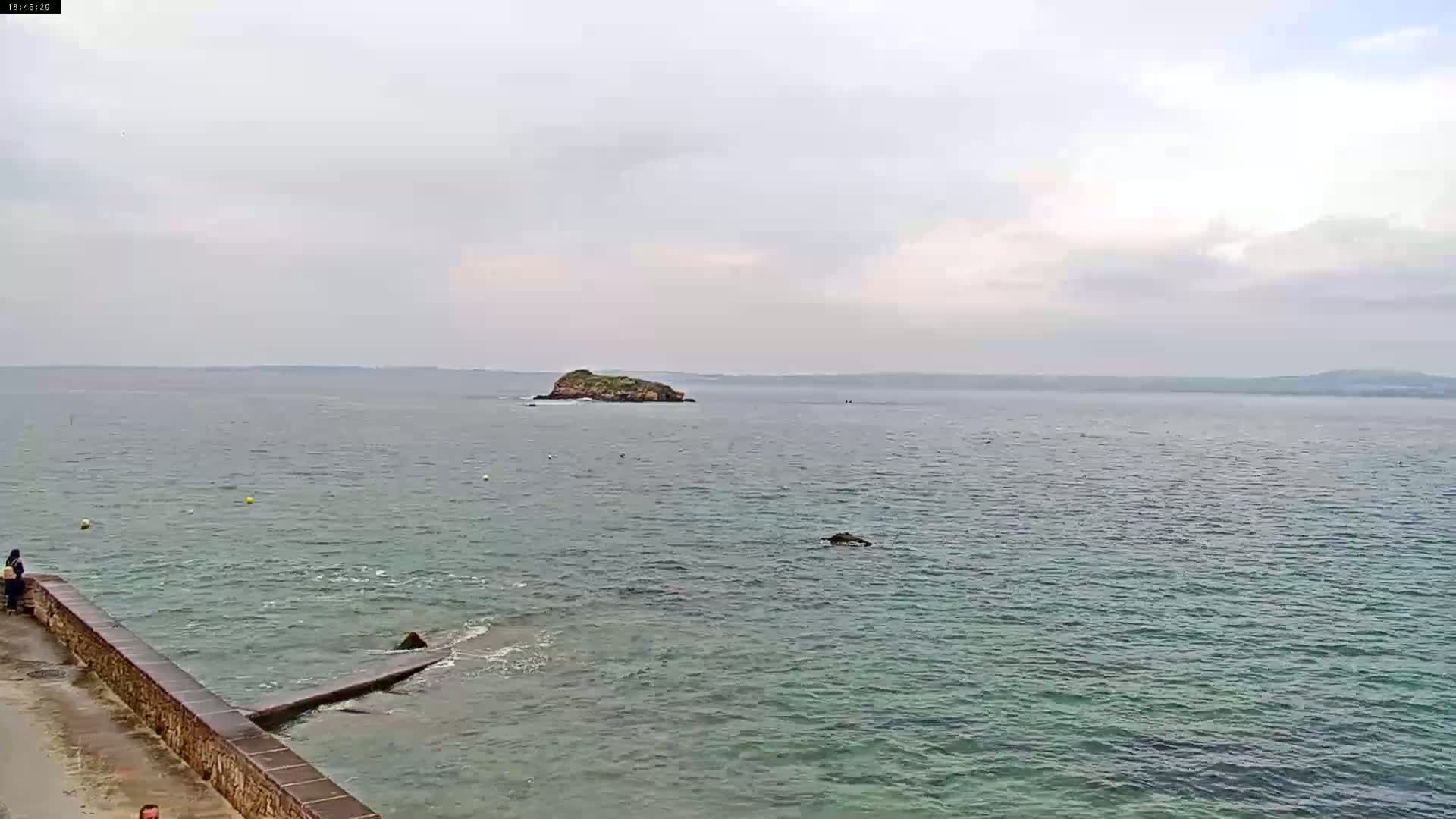 A calm, overcast day reveals a small island in the distance, viewed from a stone wall overlooking a body of water.