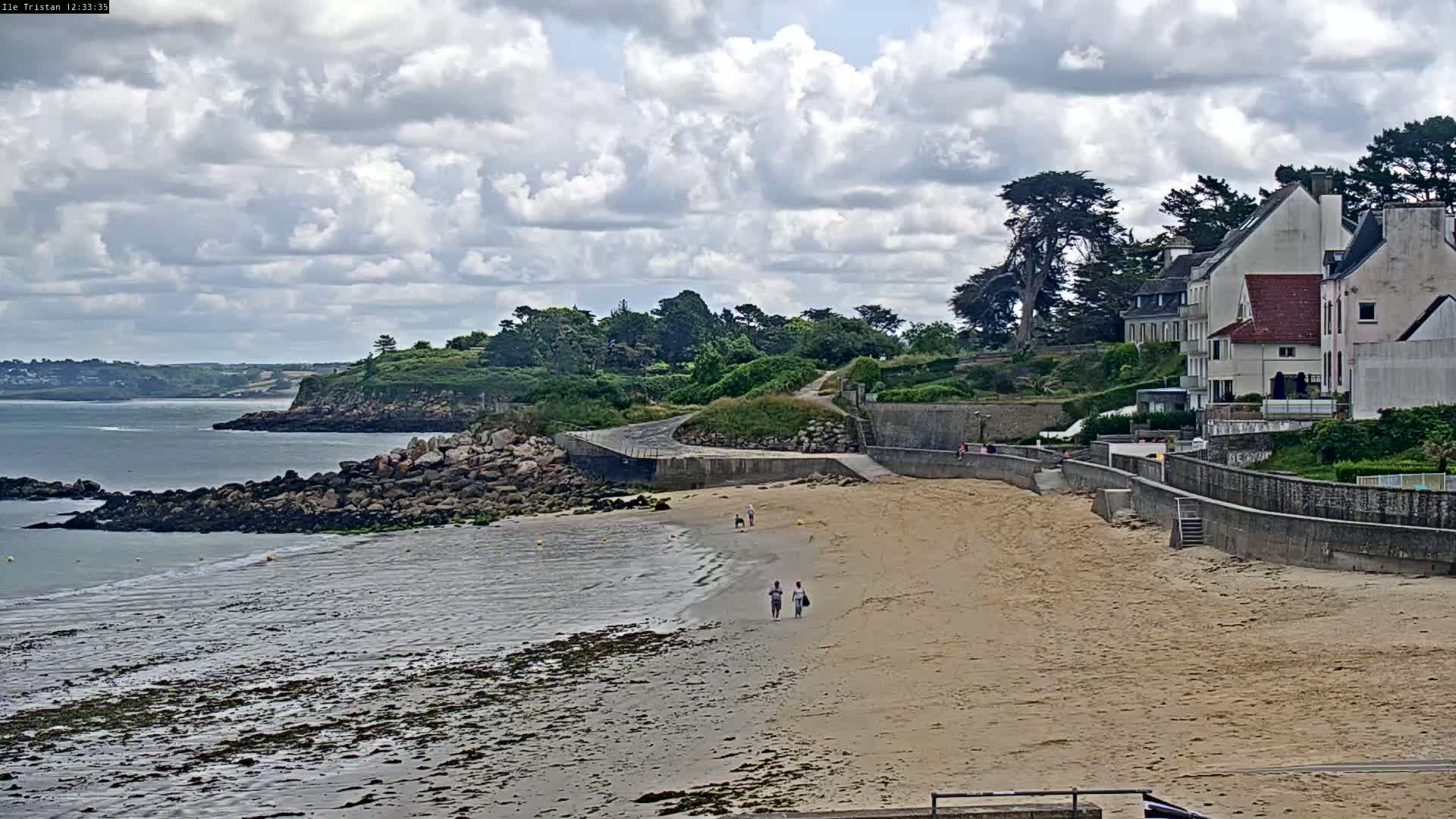 A sandy beach stretches out to a calm body of water under a partly cloudy sky, with a few people walking along the shore and houses situated on a cliff overlooking the scene.