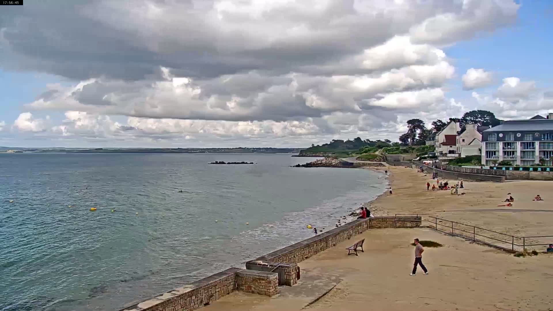 A sandy beach with a few people on it stretches along a calm body of water under a partly cloudy sky.