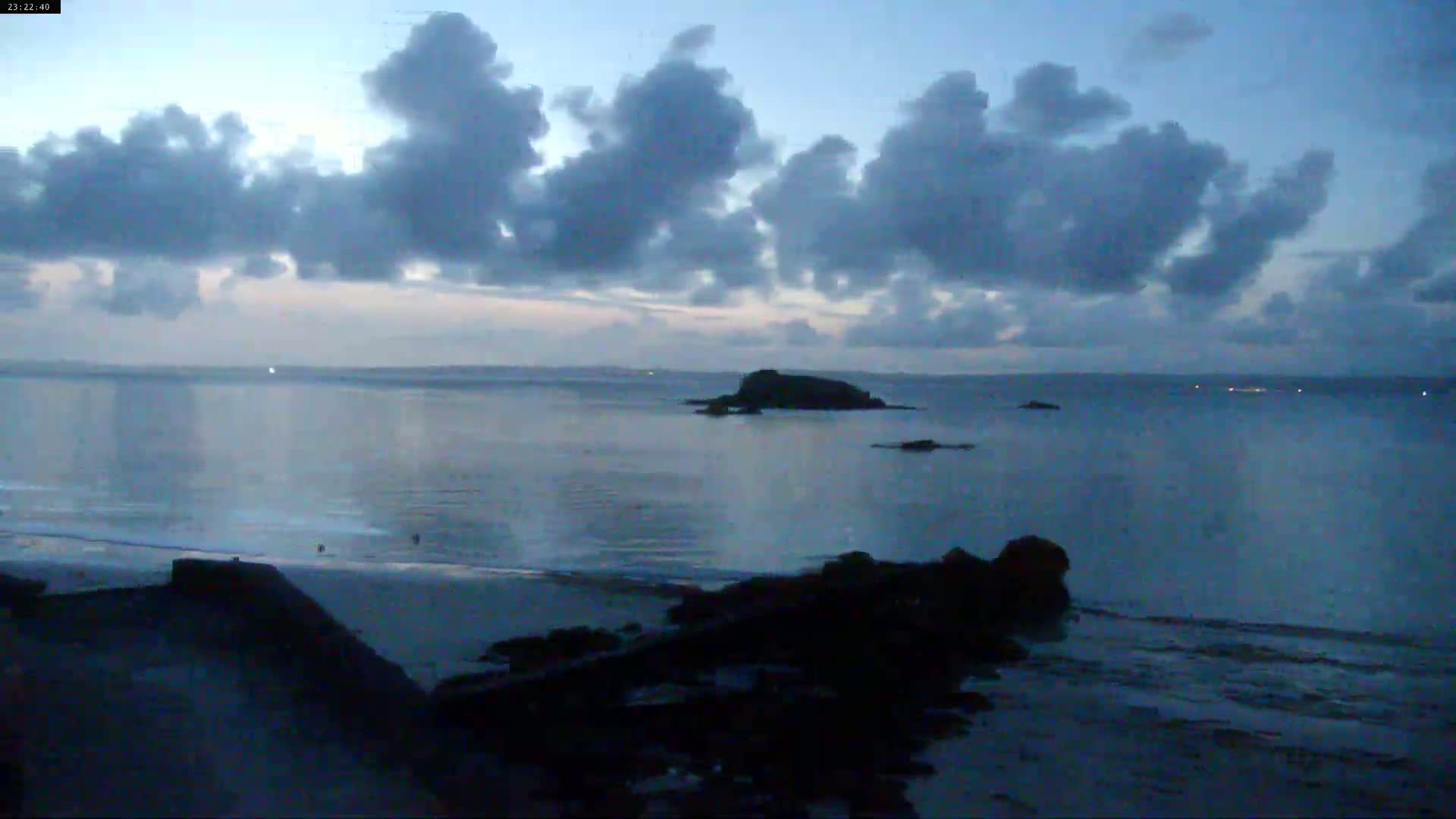 A sandy beach with a few people on it stretches along a calm body of water under a partly cloudy sky.