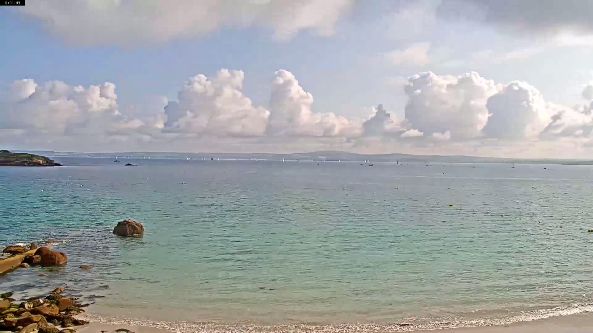 A partly cloudy day shows a calm, light teal ocean with numerous sailboats in the distance, a rocky shoreline, and a sandy beach in the foreground.