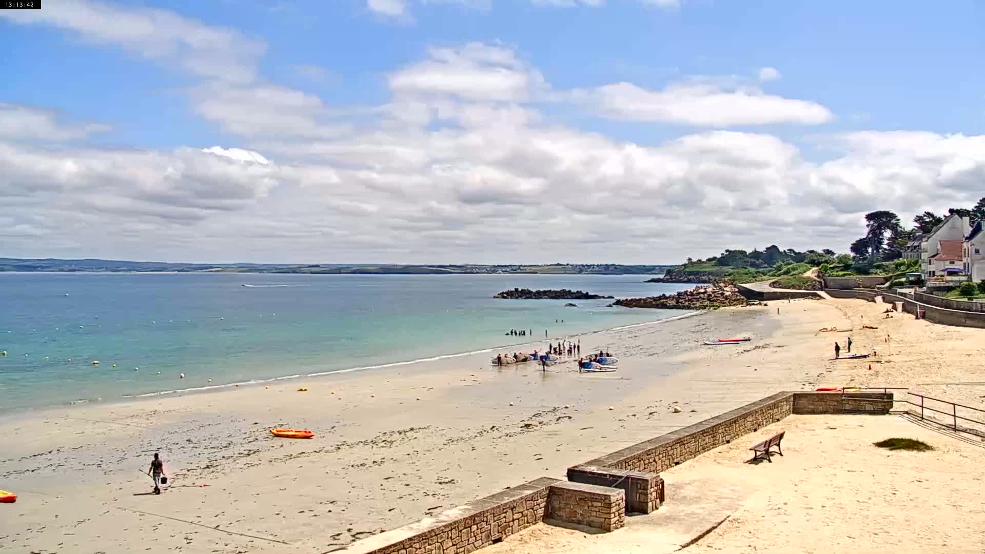 A sandy beach with calm, light-blue water stretches out under a partly cloudy sky, featuring people enjoying water activities and relaxing on the shore.