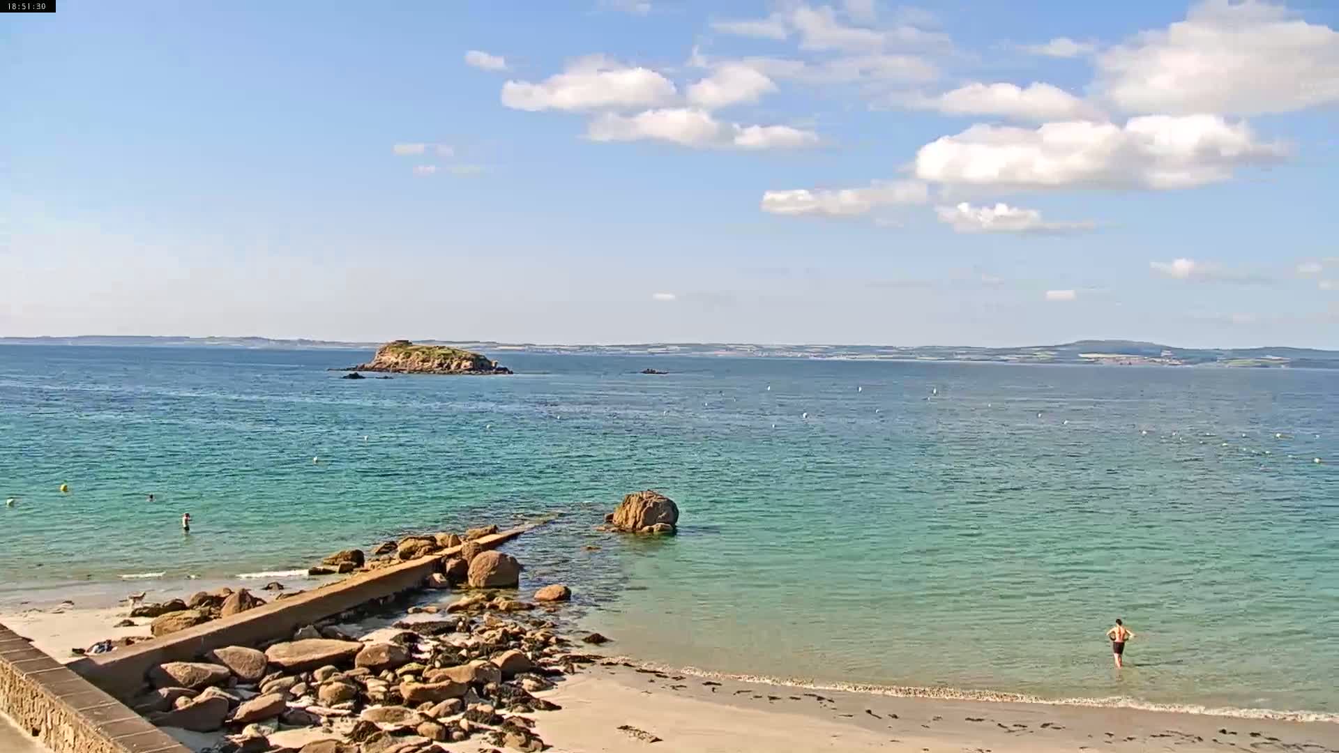 A sandy beach with a rocky shoreline and calm, clear turquoise water extends to a small island under a partly cloudy sky.