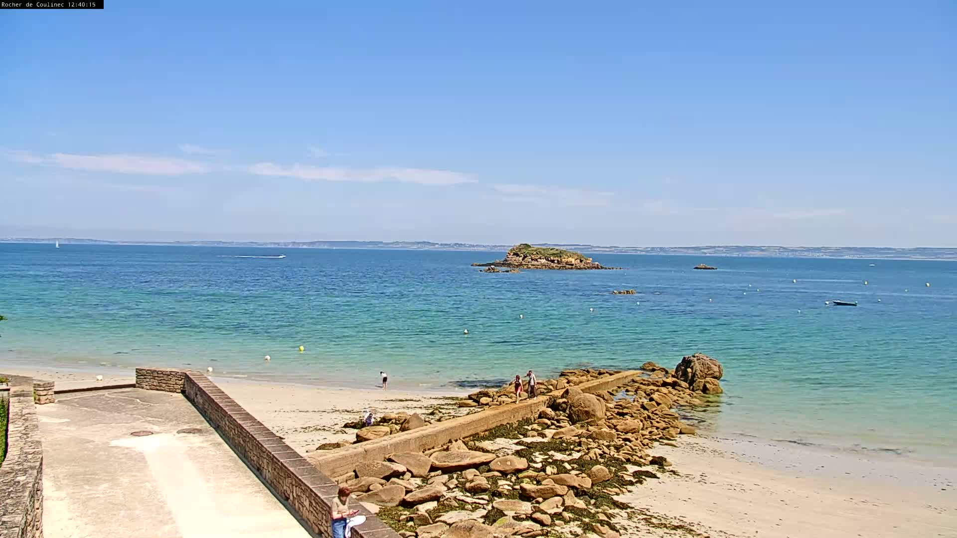A sandy beach with a rocky shoreline and a small island in the distance under a clear, sunny sky.